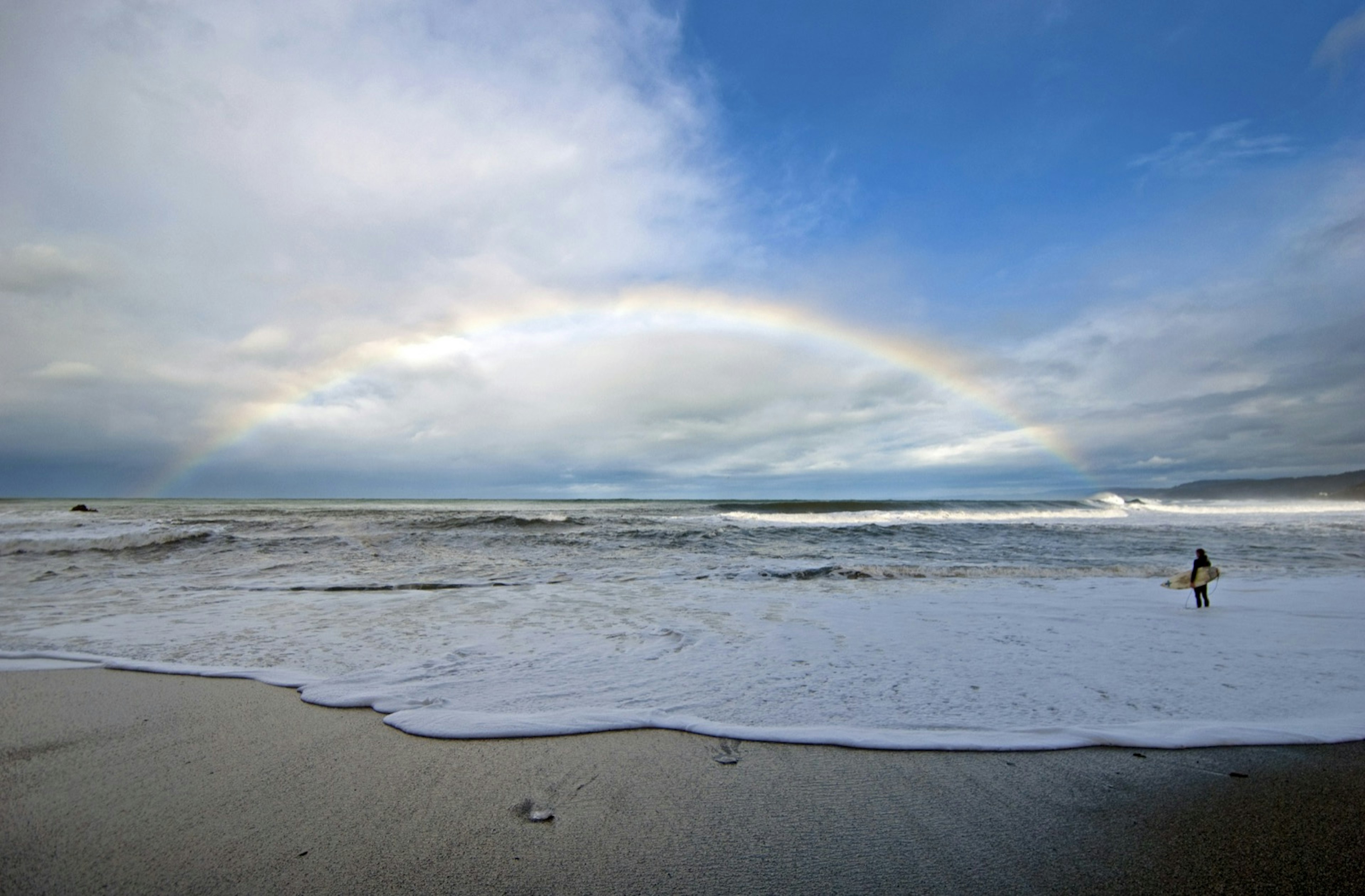 A surfer stands in the foam looking out at a rainbow on the horizon