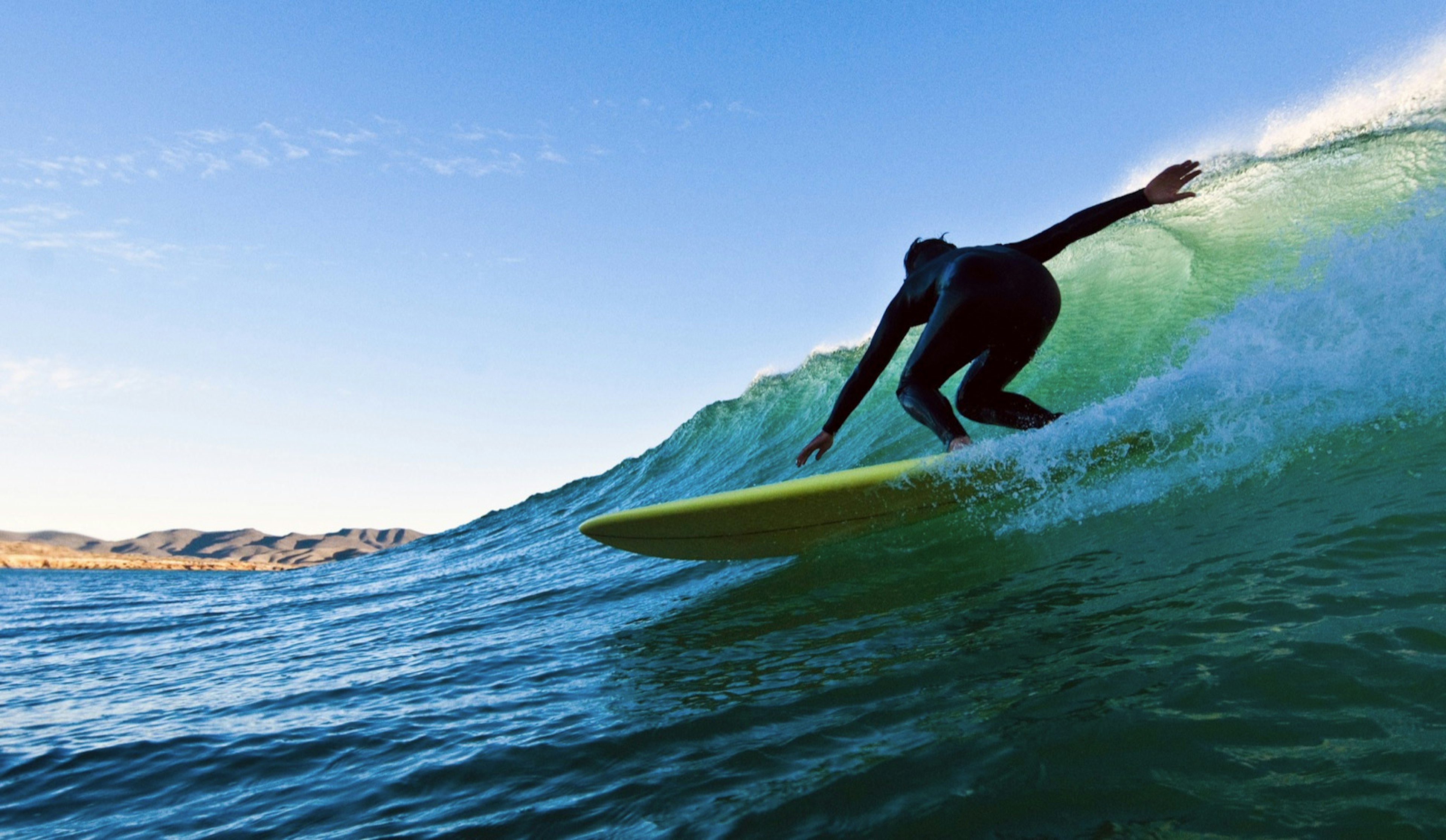 A surfer in a black wetsuit slides down a green wave with the sun behind it in Mexico