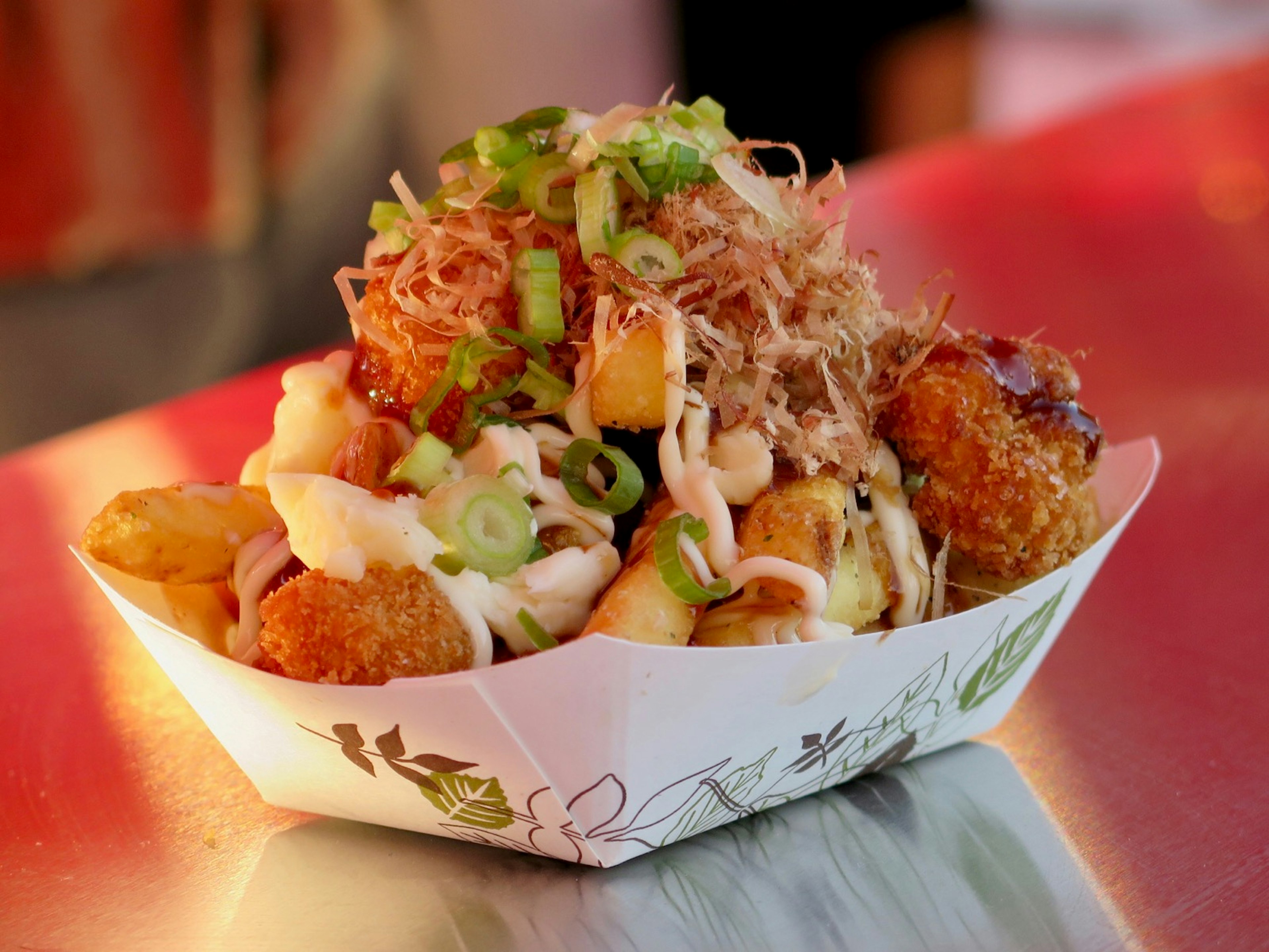 A large pile of fries, with gravy and chives and other various toppings is seen in a paper boat on a table