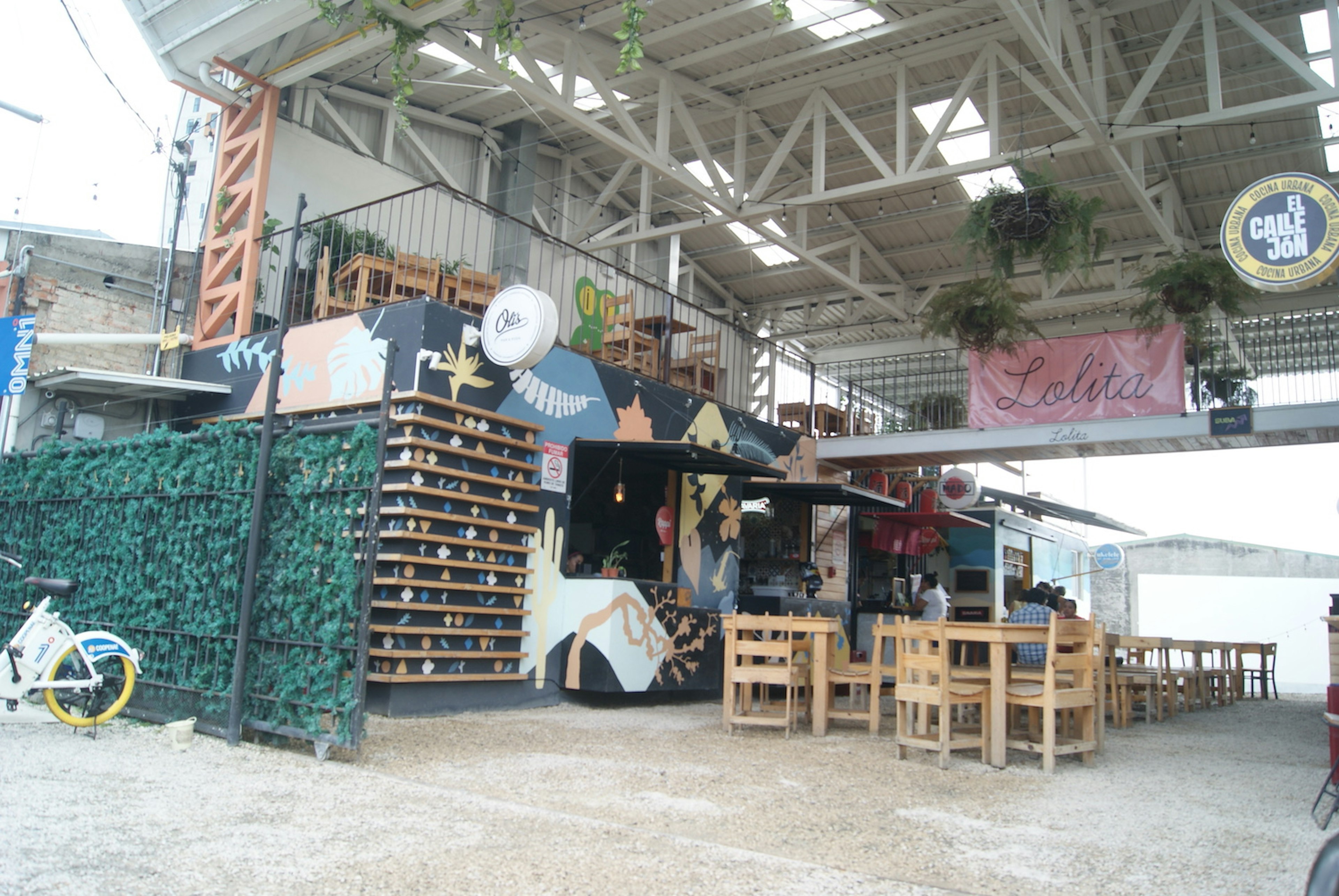 Exterior view of food stalls at Jardin de Lolita. Large plants and lights hang from the exposed rafters.