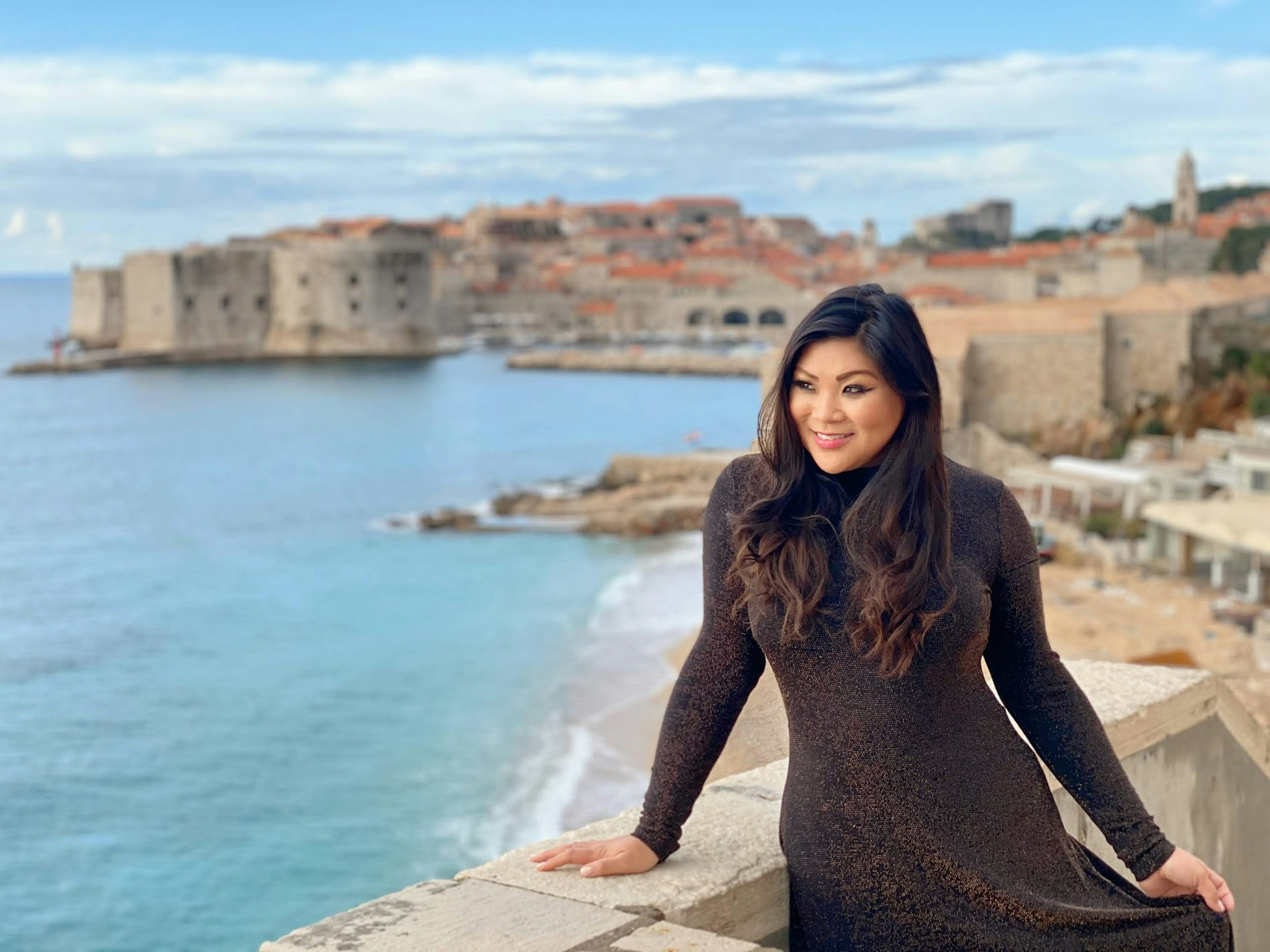 Woman stands next to a coastal wall, with old structures in background