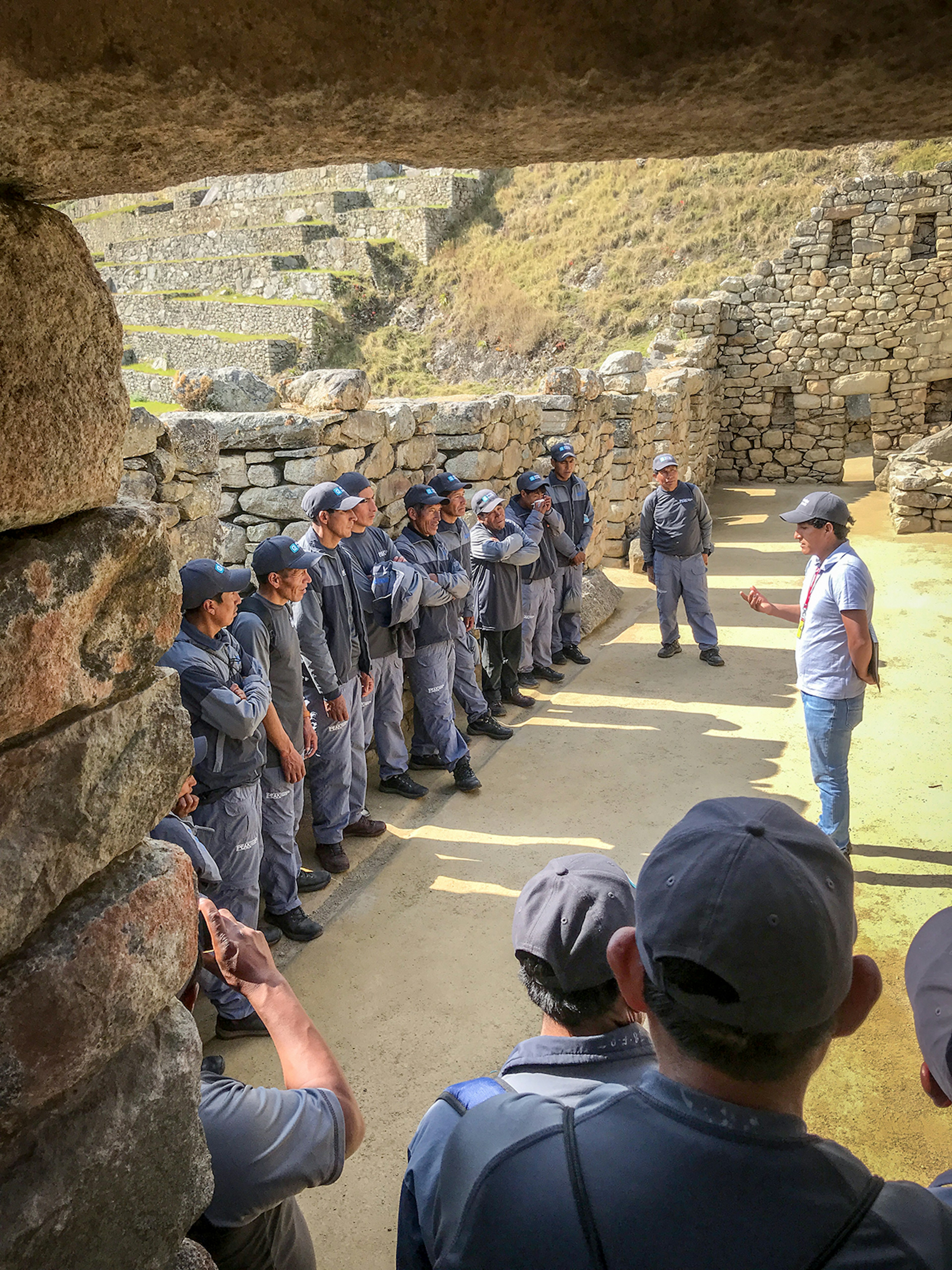 Men stand along a stone wall at Machu Picchu listening to a guide speak about the site