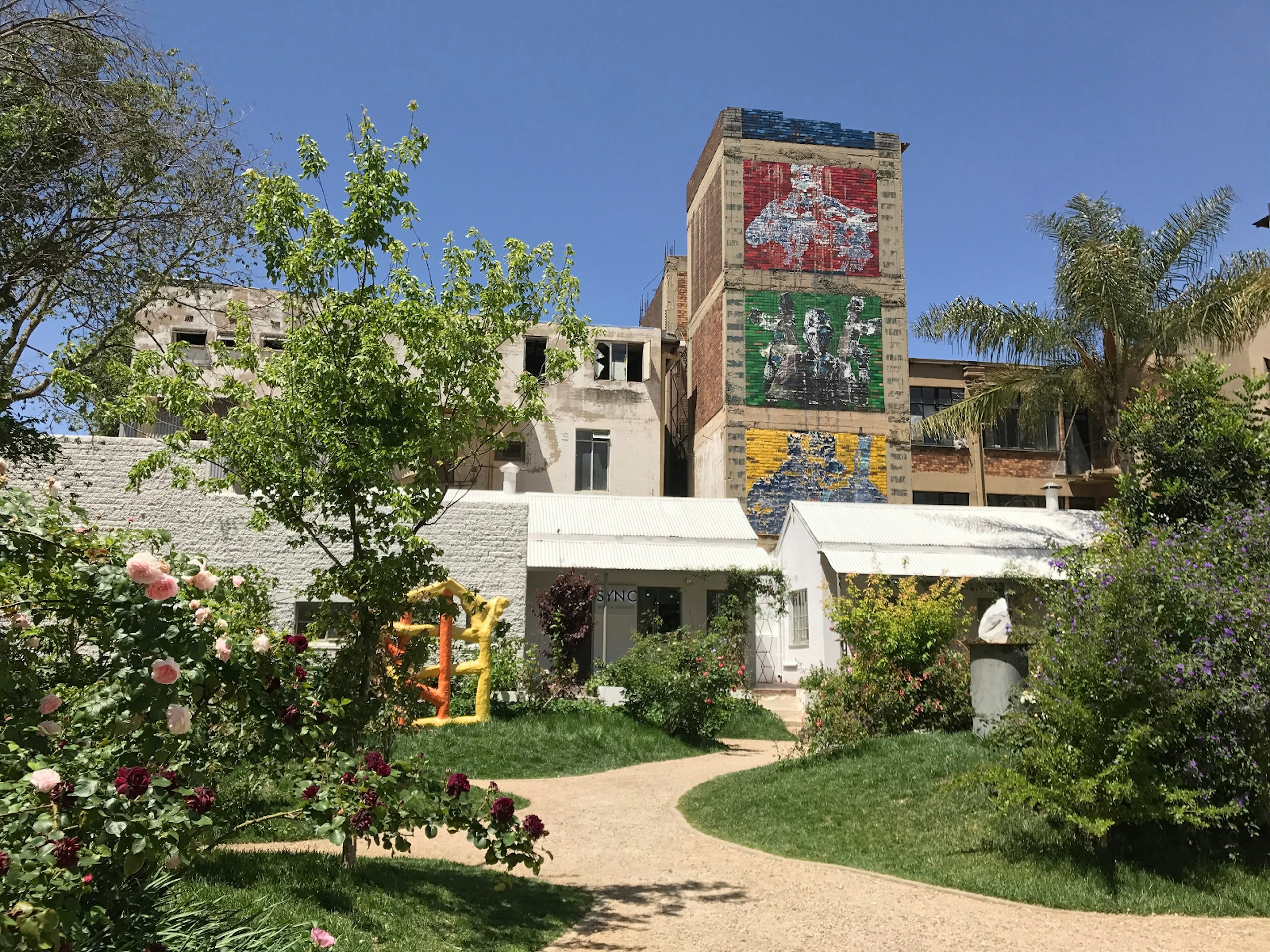 A gravel path wynds between grass and trees, with the colorful, mural-clad old warehouse behind © Simon Richmond / Lonely Planet