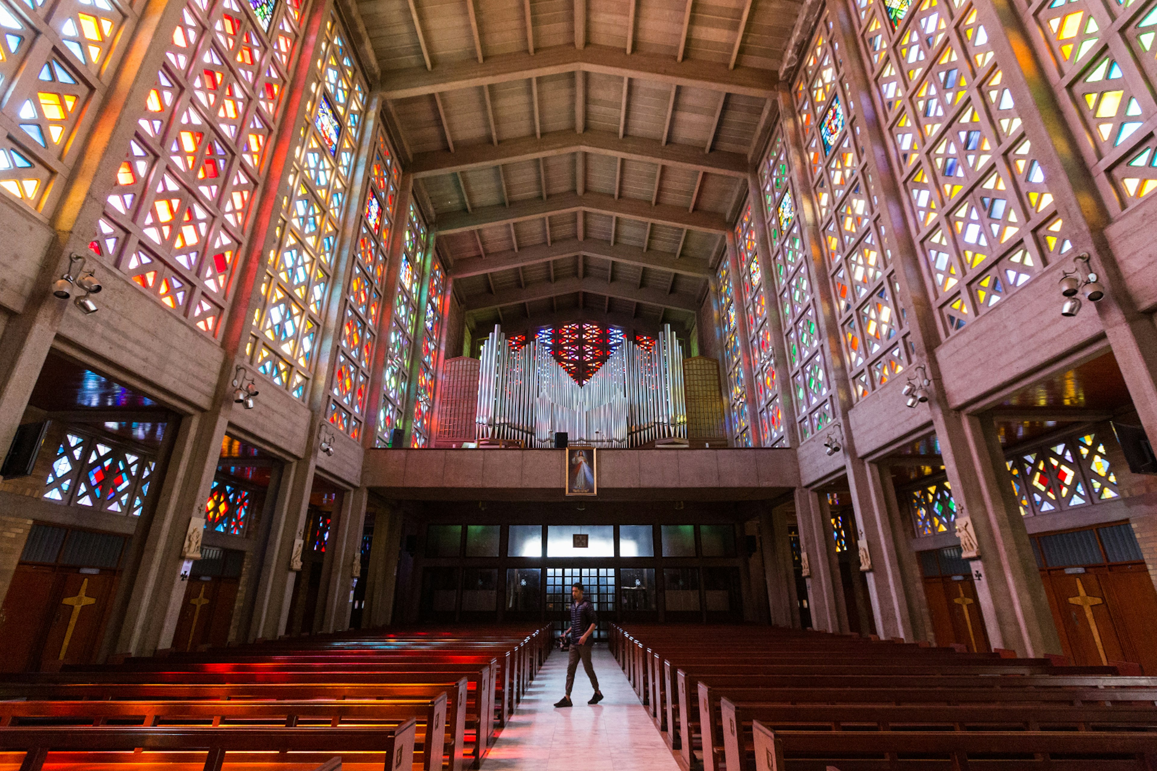 Looking down the long aisle of the Cathedral of Christ the King in Hillbrow. The towering vertical walls to the left and right are brilliantly lit stained glass windows, above is a modern vaulted ceiling.