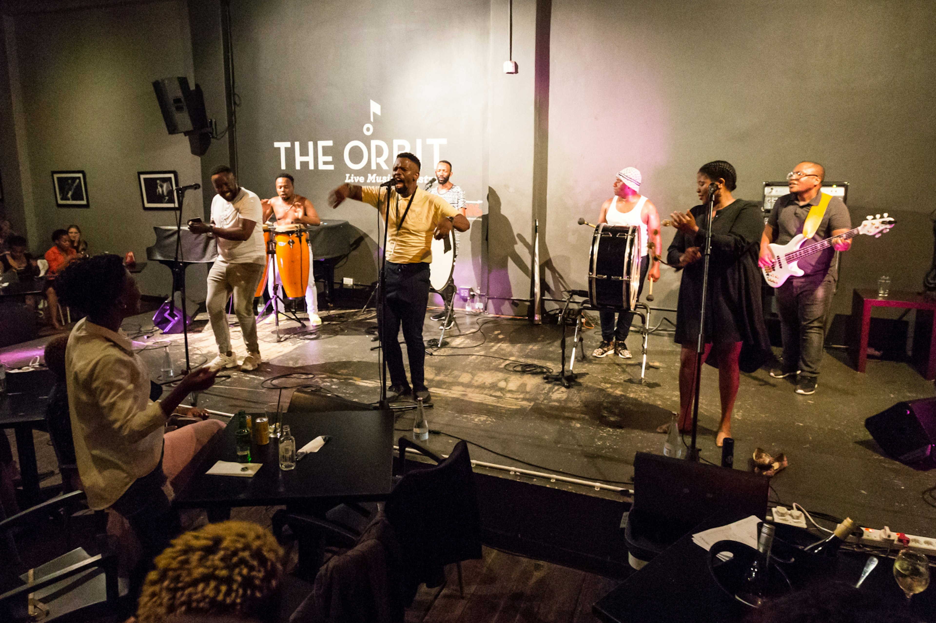 A singer belts out jazz vocals, backed by three different drummers, a guitarist, percussionist and back-up singer in the Orbit jazz club. Guests sitting around raised tables enthusiastically watch on.