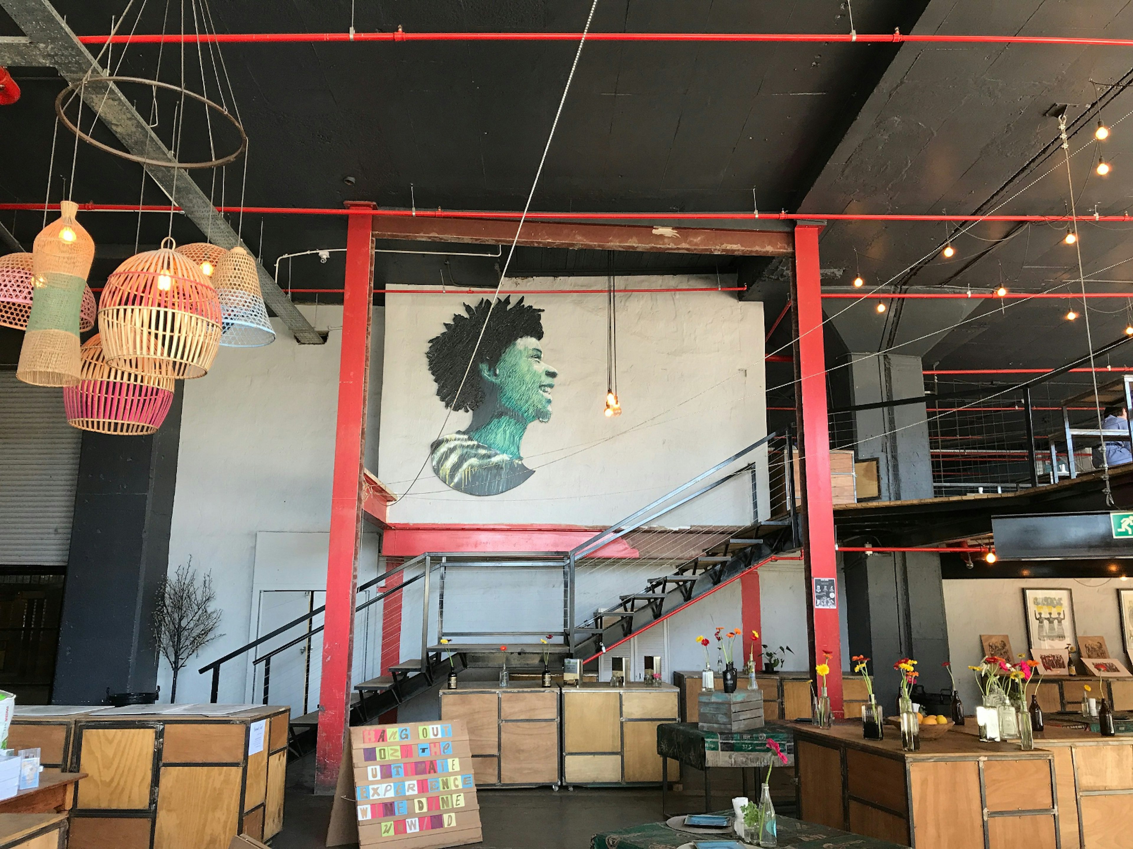 Beneath the black ceiling of this large warehouse are bright red steel support beams and sprinkler pipes. A portrait of a smiling woman has been painted on the wall, colourful wicker lanterns hang in a group and simple wooden cabinets are scattered around, topped with vases of flowers, framed pictuers and artworks. © Simon Richmond / Lonely Planet