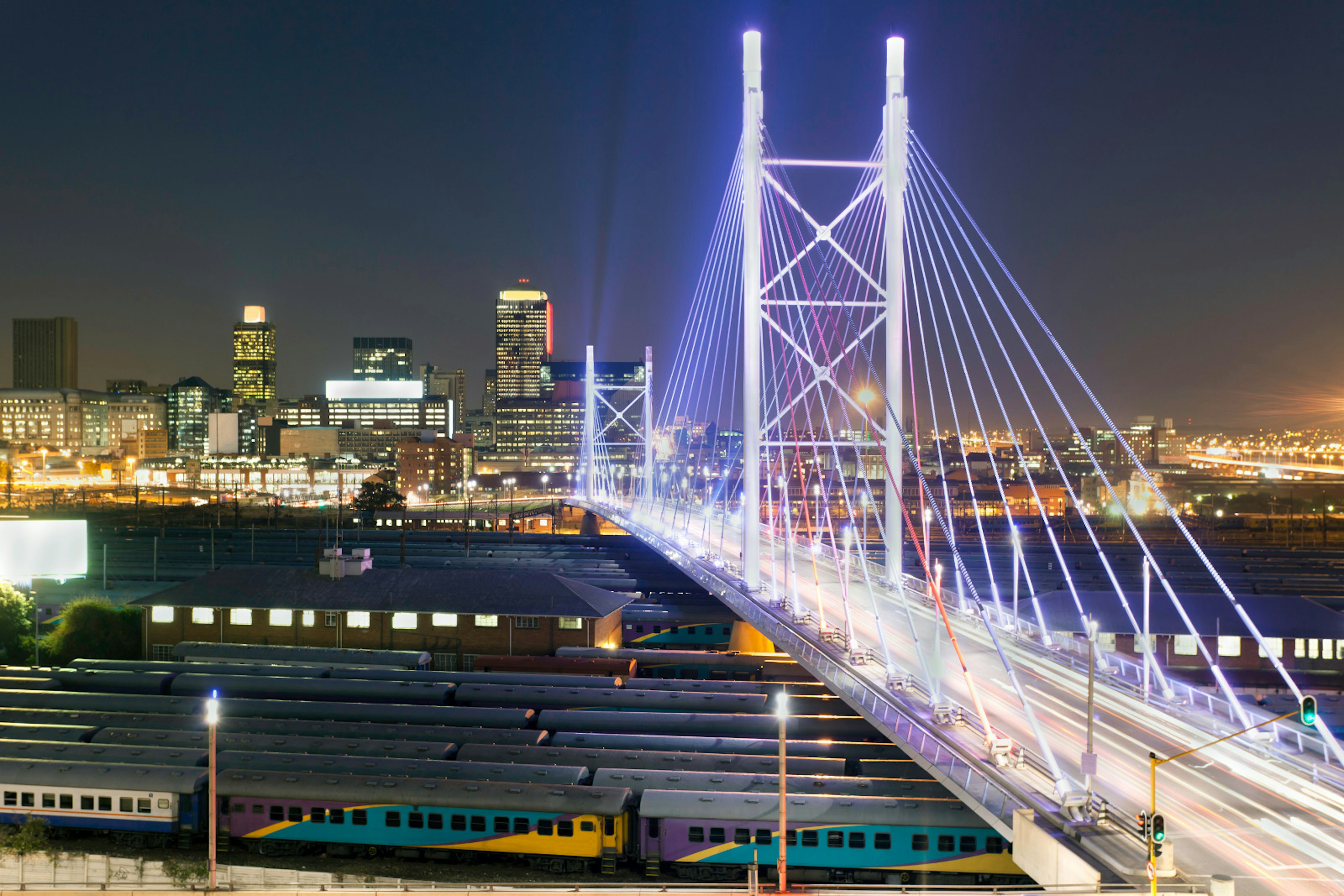 Stretching over eight rows of parked trains, the brilliantly lit white towers and cables of the Nelson Mandela Bridge stretch towards the skyline © Henrique NDR Martins / Getty Images