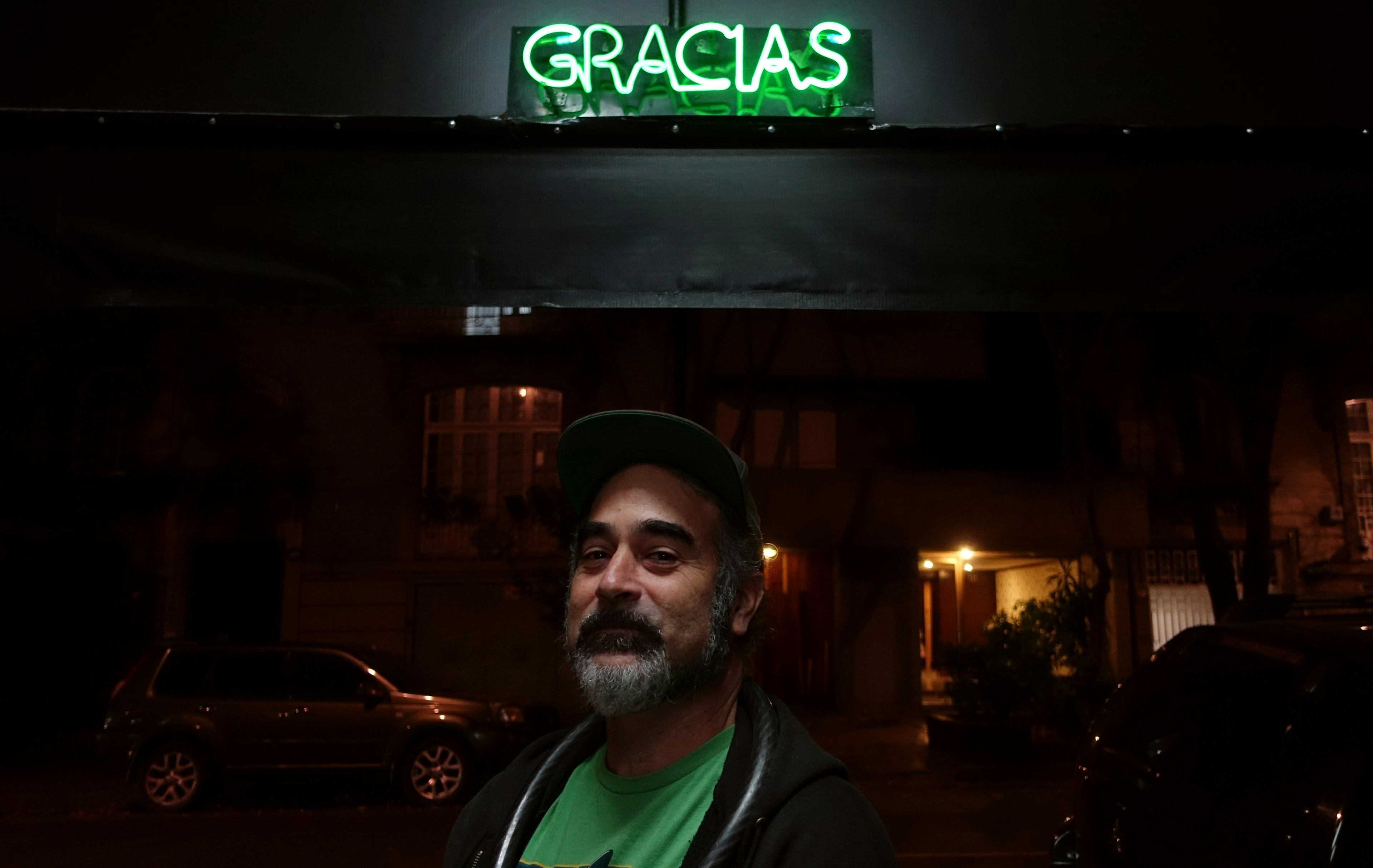 A man wearing a dark baseball hat, green t-shirt and black sweatshirt stands under a neon sign that says