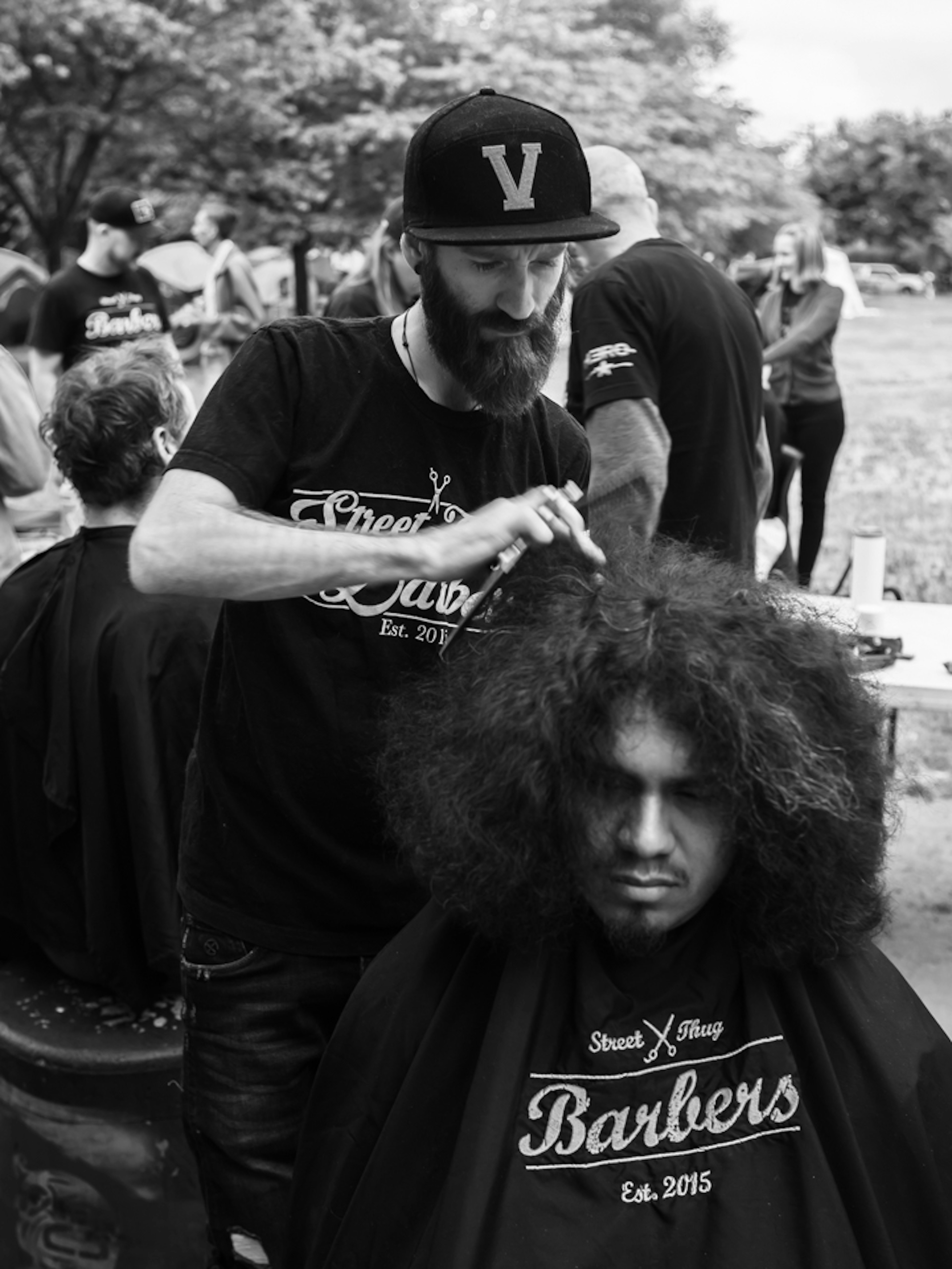 A bearded man in a flatcap is giving a man with long hair a haircu.