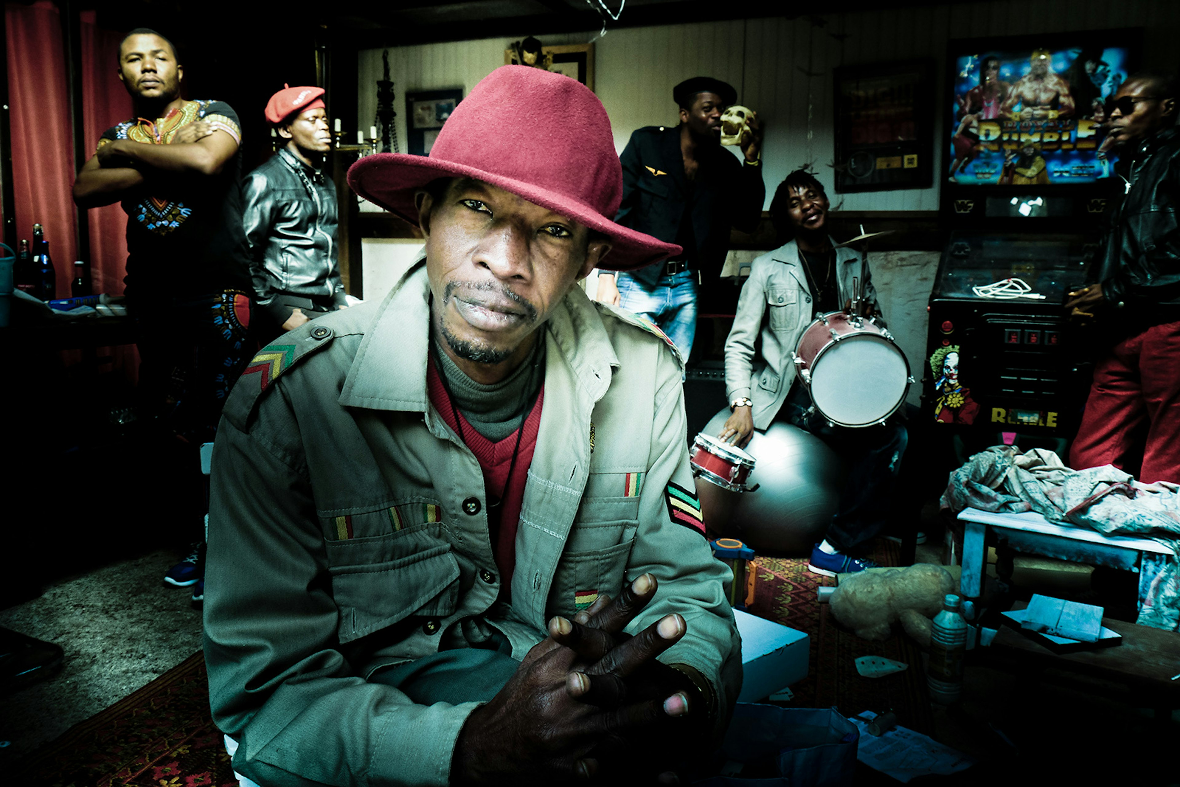 A man wearing a wide-brimmed red hat sits close to the camera, surrounded by his bandmates in various poses