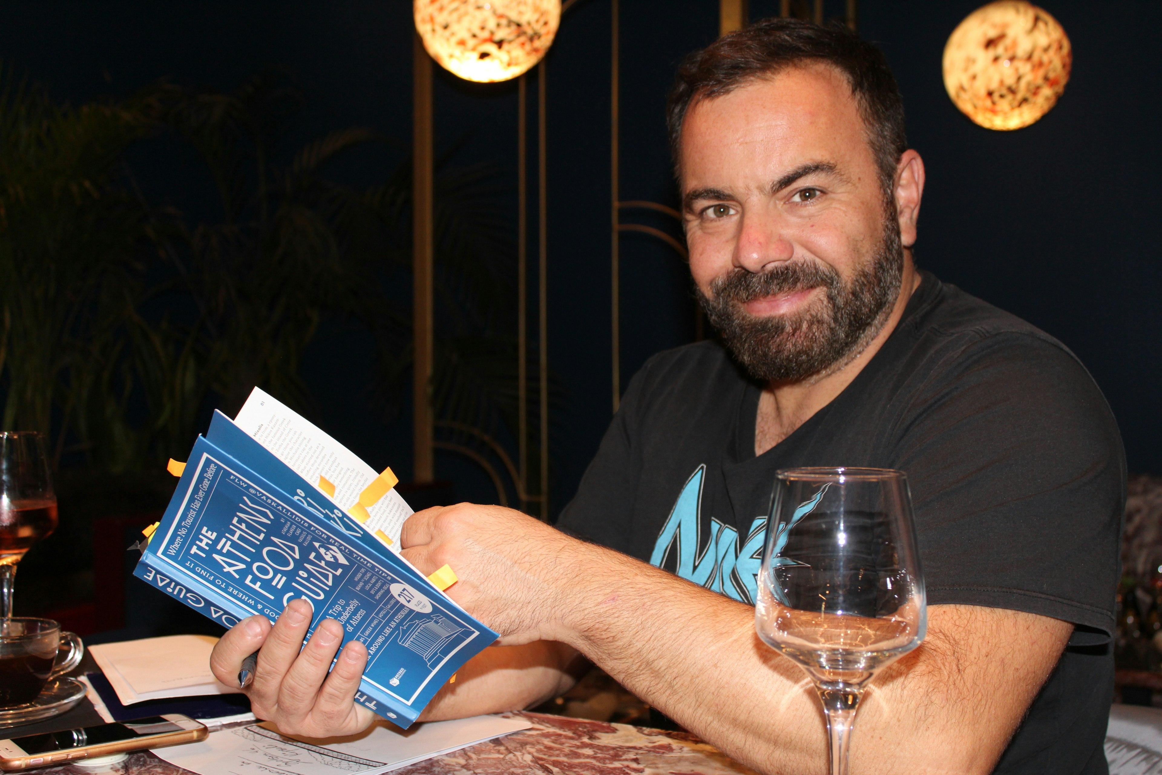 Vassilis Kallidis smiles confidently at the camera as he holds a copy of his new book, the Athens Food Guide under the warm lighting of blown glass lamps hanging from the ceiling of a restaurant. He wears a black t-shirt and a wine glass sits in front of him. He has a short beard and dark, short-cropped hair.