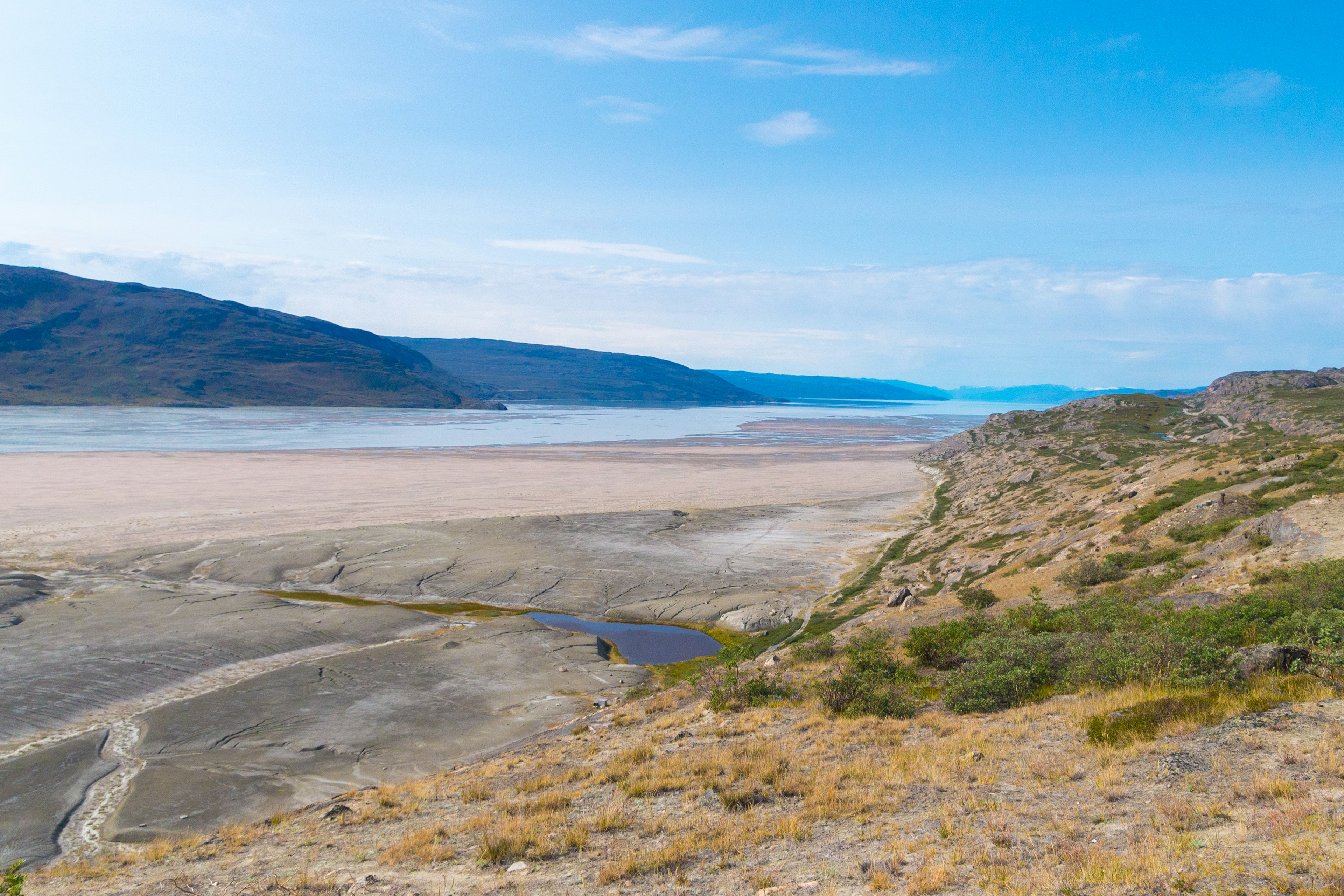 A view over a sandy beach to the waters of Kangerlussuaq fjord, with gently sloping slopes on the opposite side