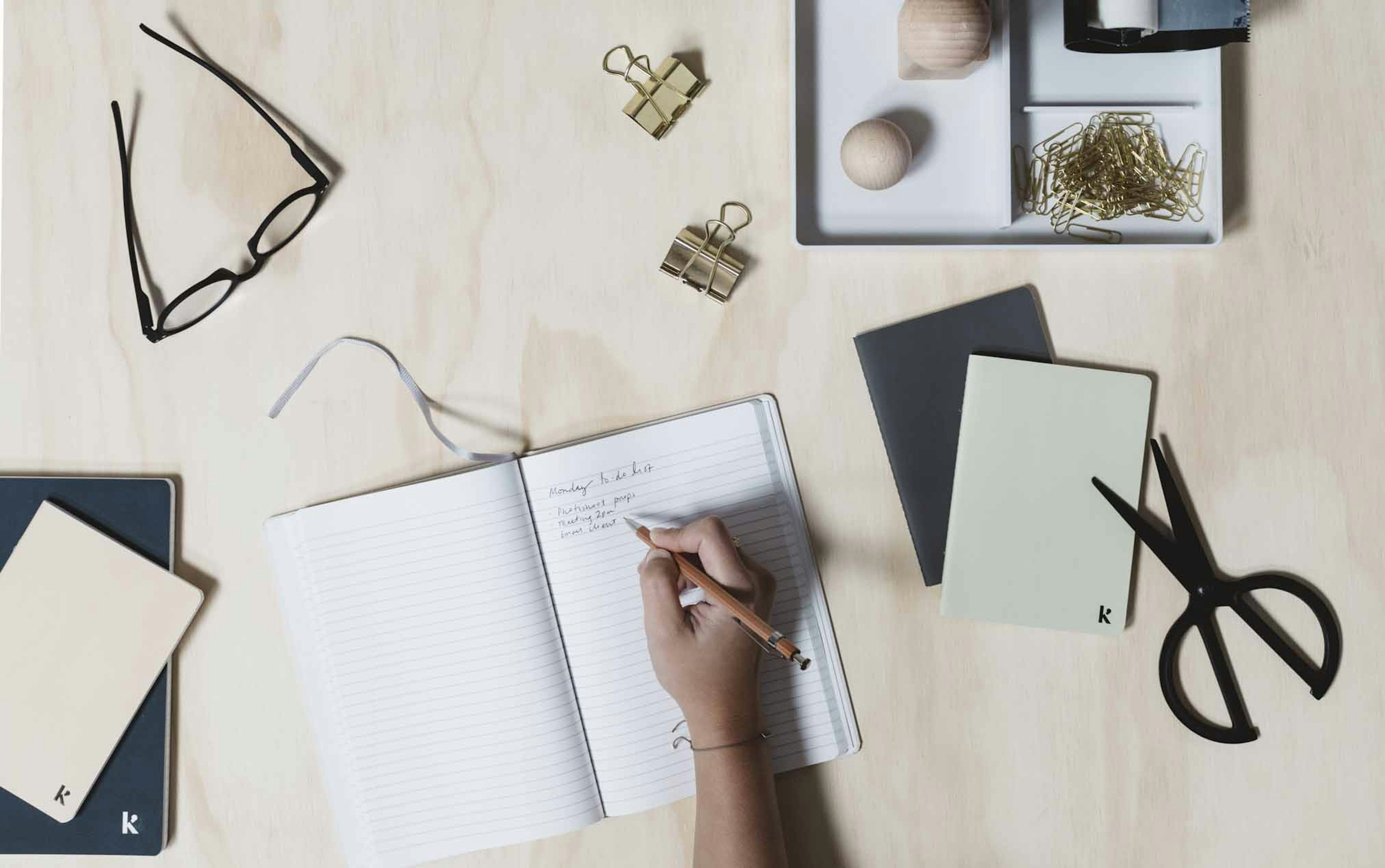 A desk with scissors, paper clips, and a hand writing in an open notebook