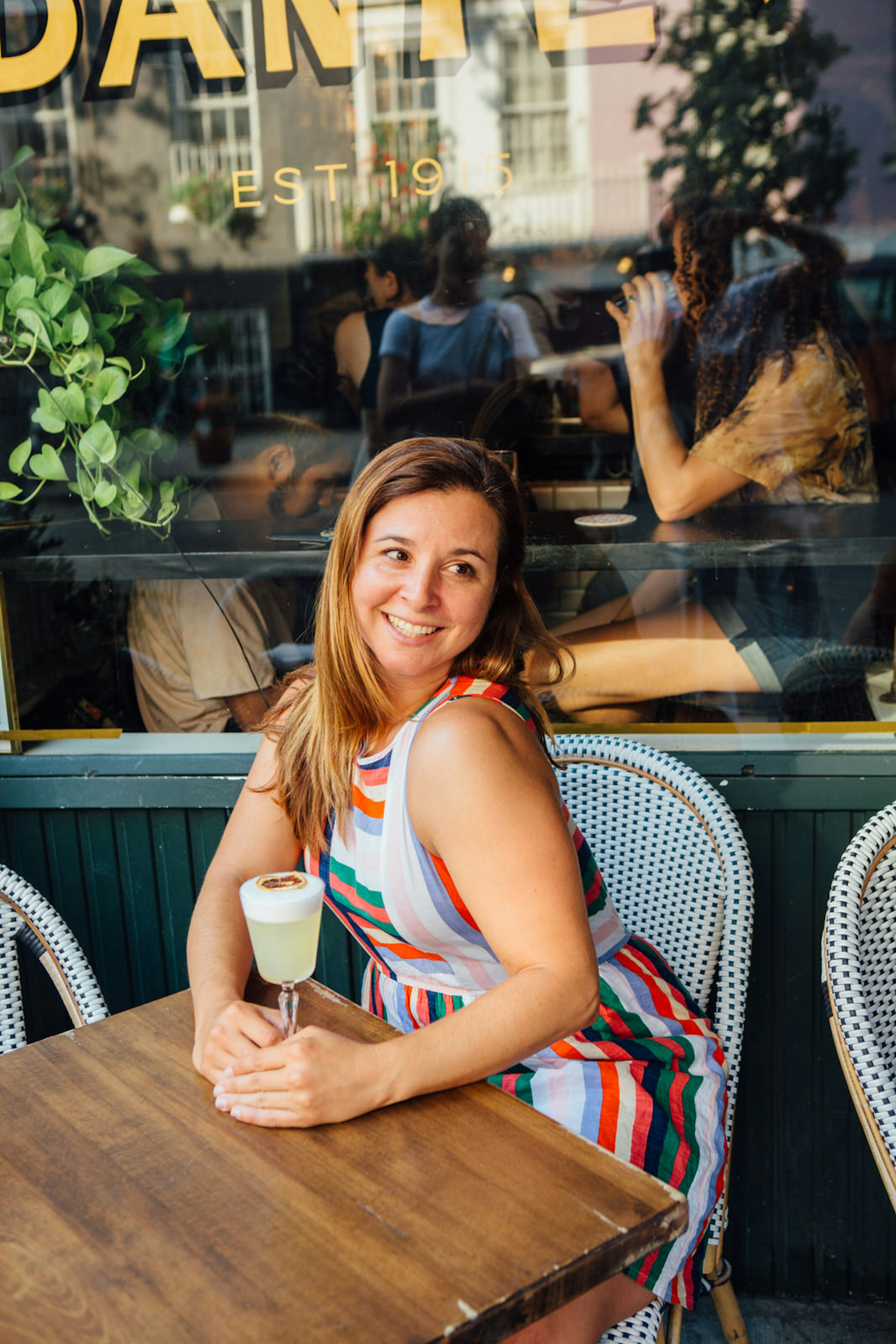 A woman smiling, sitting outside a restaurant and looking over her shoulder away from the camera. She is wearing a stripy dress and has her hand on a glass of wine on the table