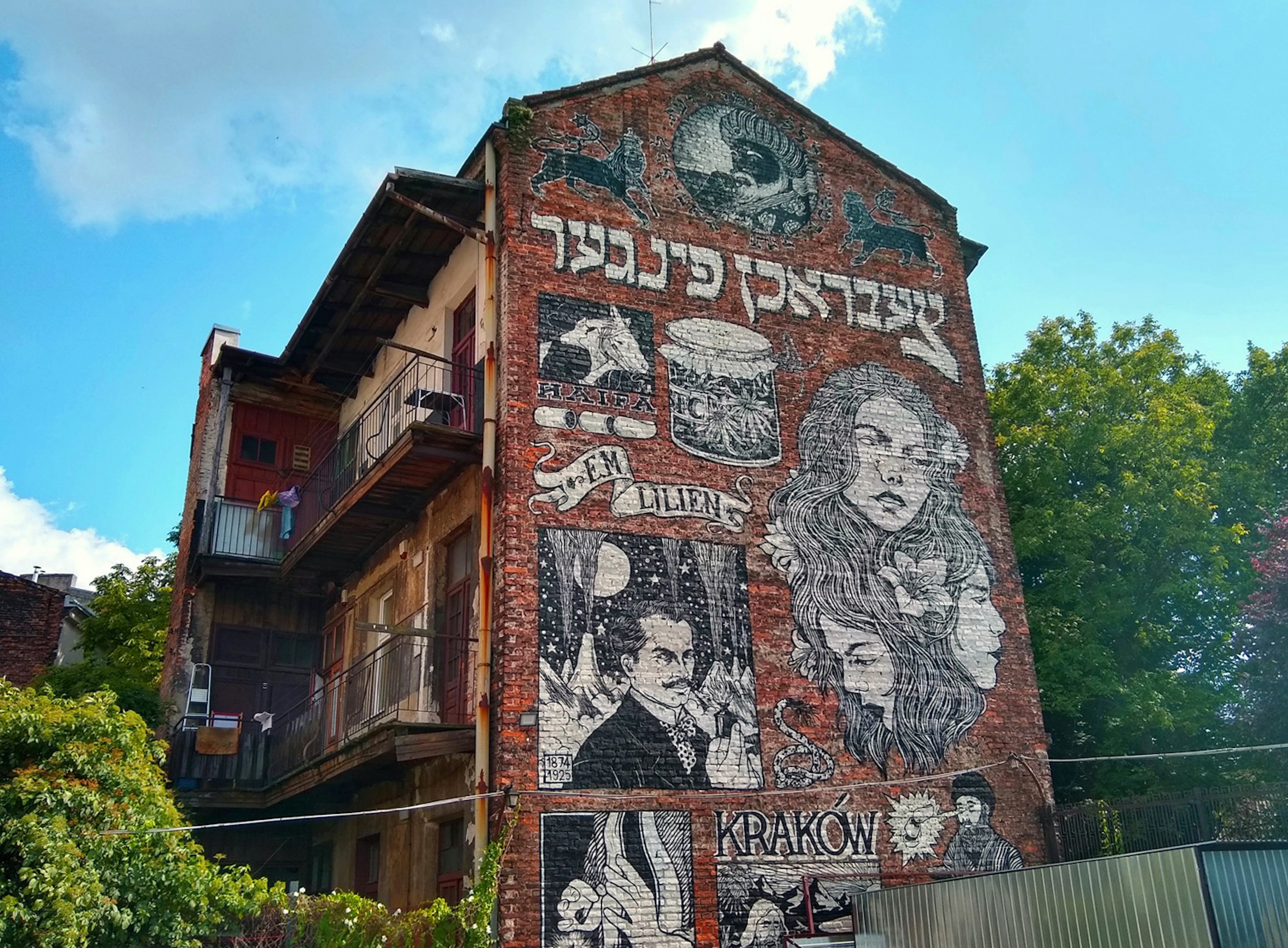 The side of a red brick three-story building shows a black and white mural with Hebrew writing and the prominent image of a woman; Krakow Jewish cultural revival