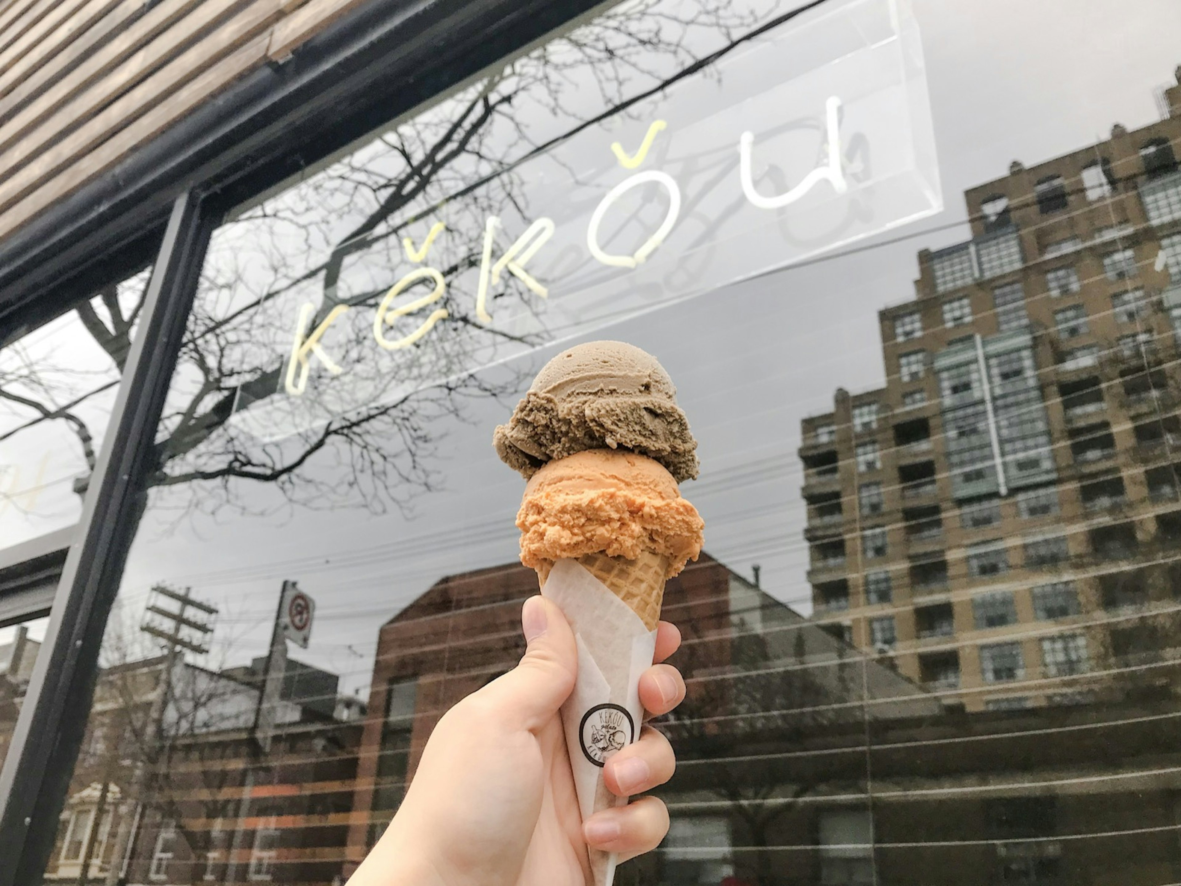 The city of toronto is reflected in a shop window as a hand holds an ice cream cone with two earth-toned scoops