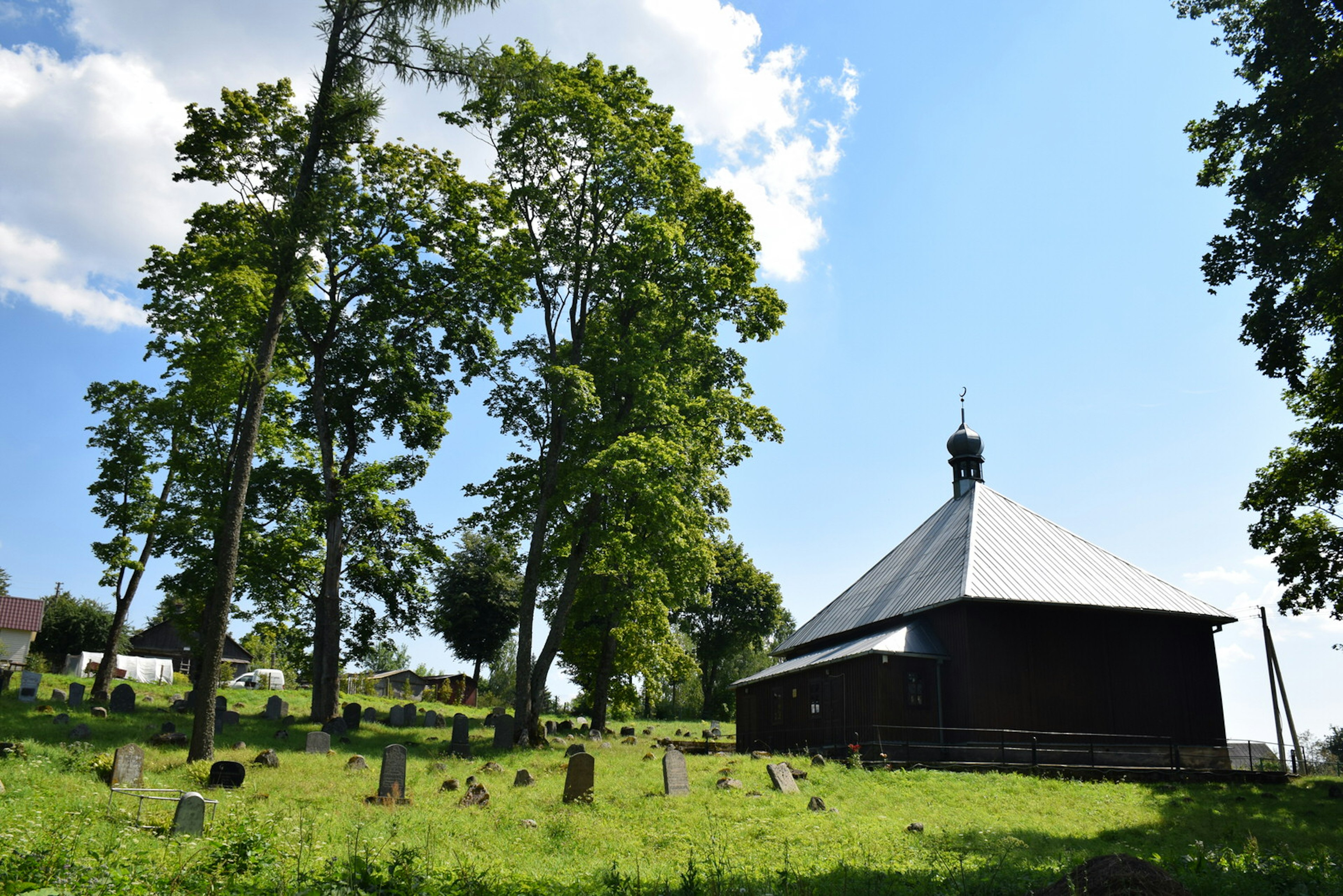 A simple, square, dark wood mosque stands with very tall trees on each side, with a cemetery in the foreground.