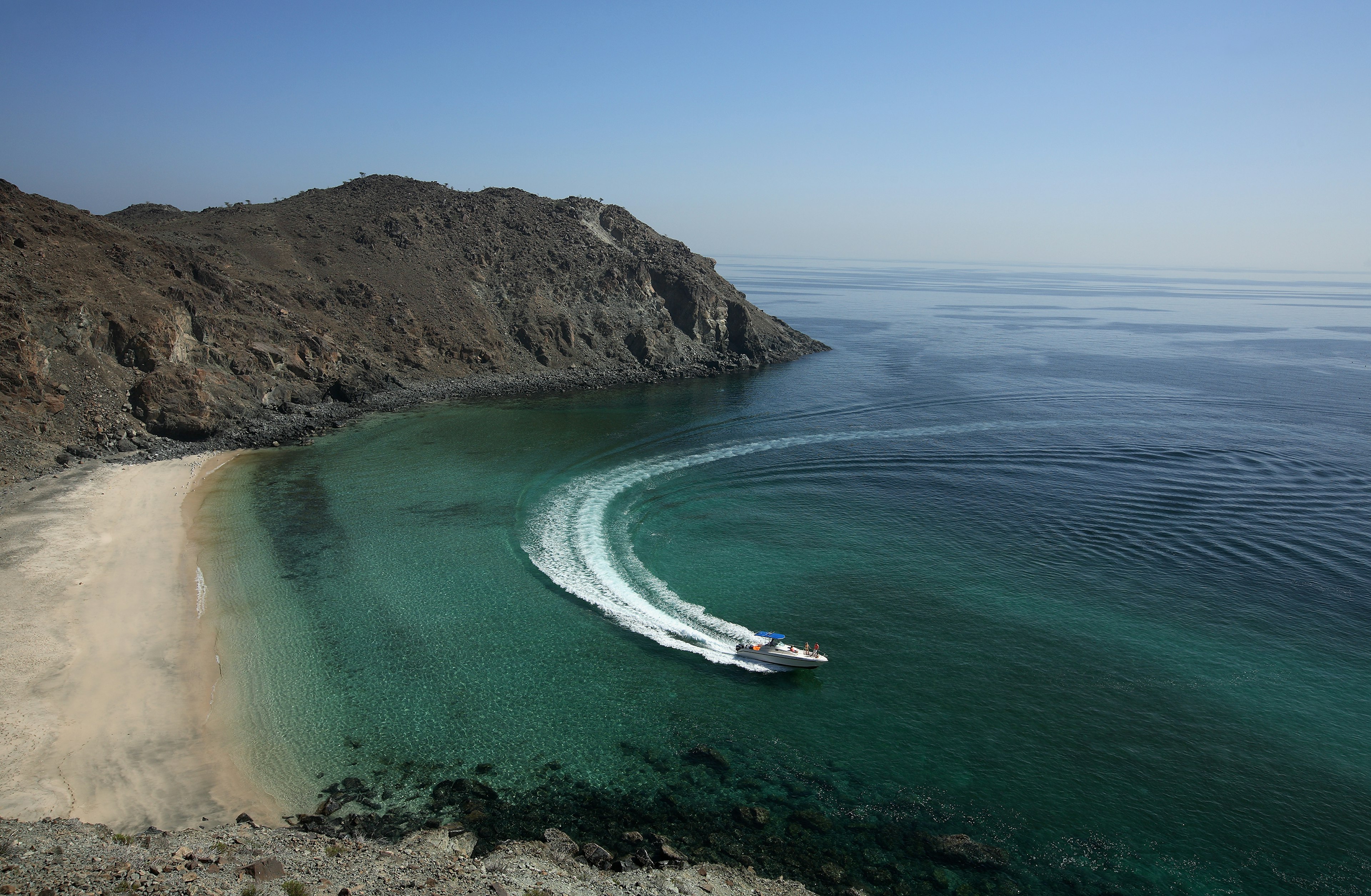 A speed boat cuts a sharp turn through the water at a sandy crescent beach that is hemmed in by dark rocky outcrops.