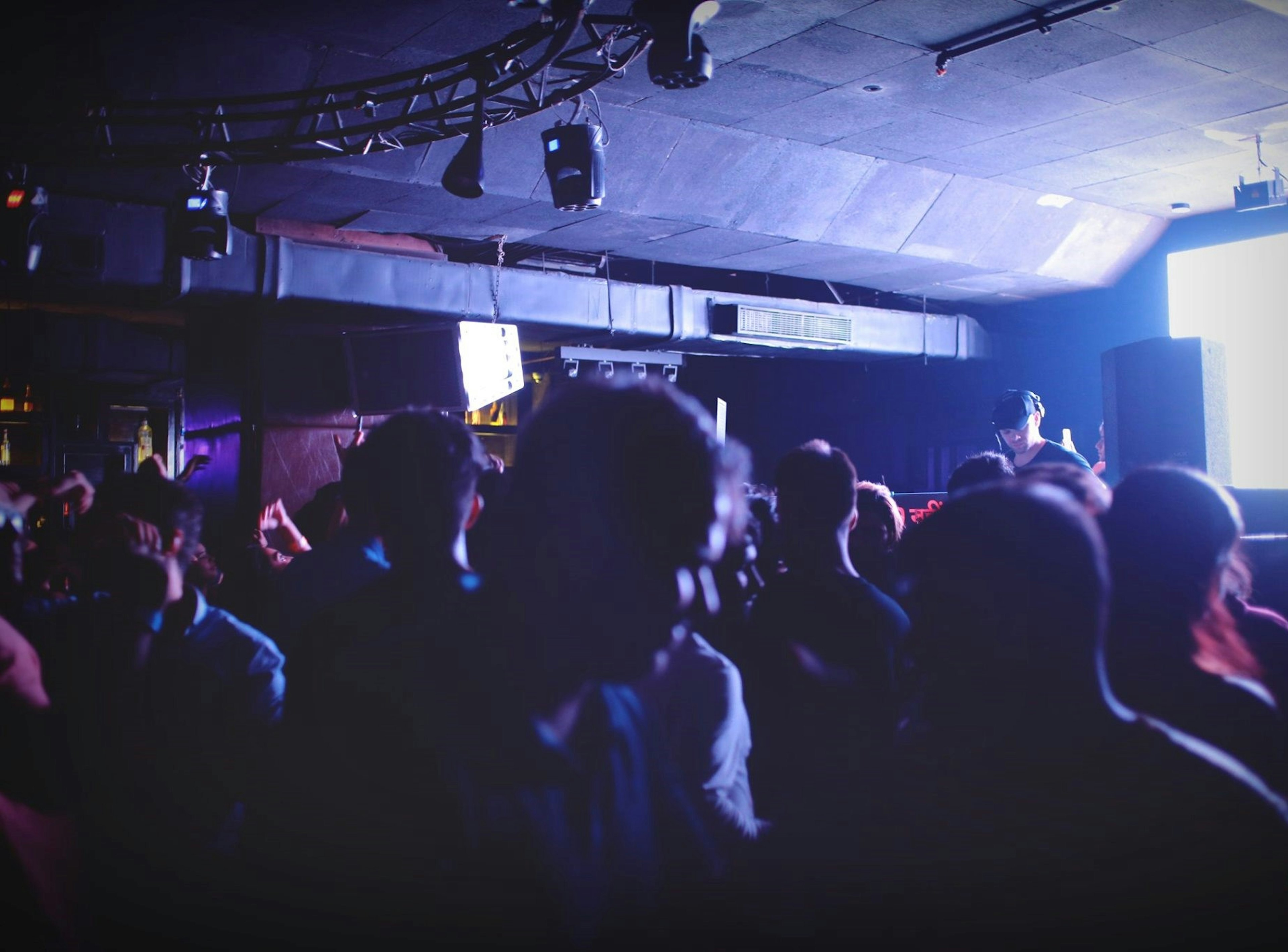 A view of the interior of a nightclub. The dance floor, filled with people, is dark and in shadow, while the DJ booth, where a man stands at the decks, is lit up.