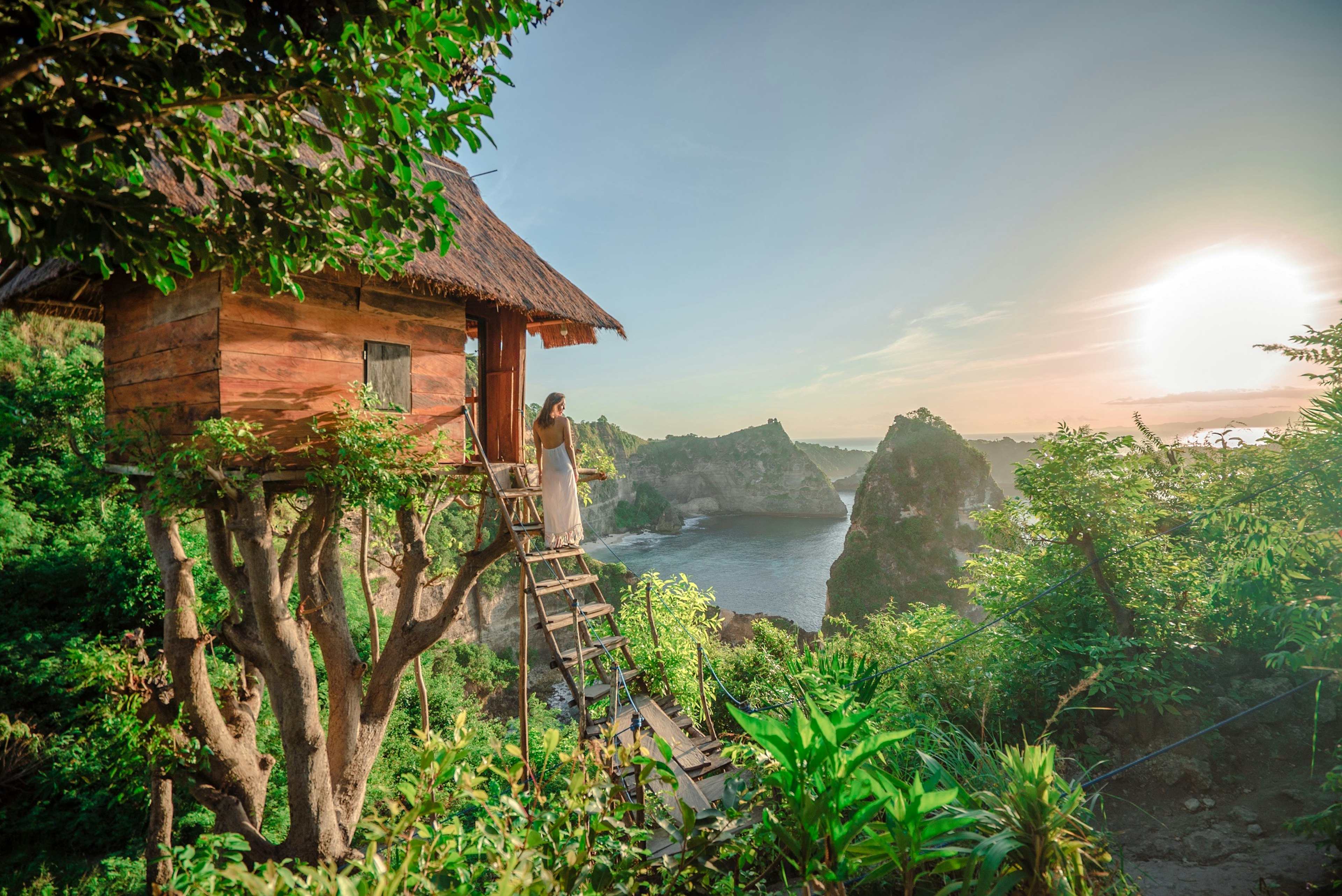 A woman in a long white flowing dress stands on the steps to a wooden tree house looking out over rocky outcrops down to the sea.