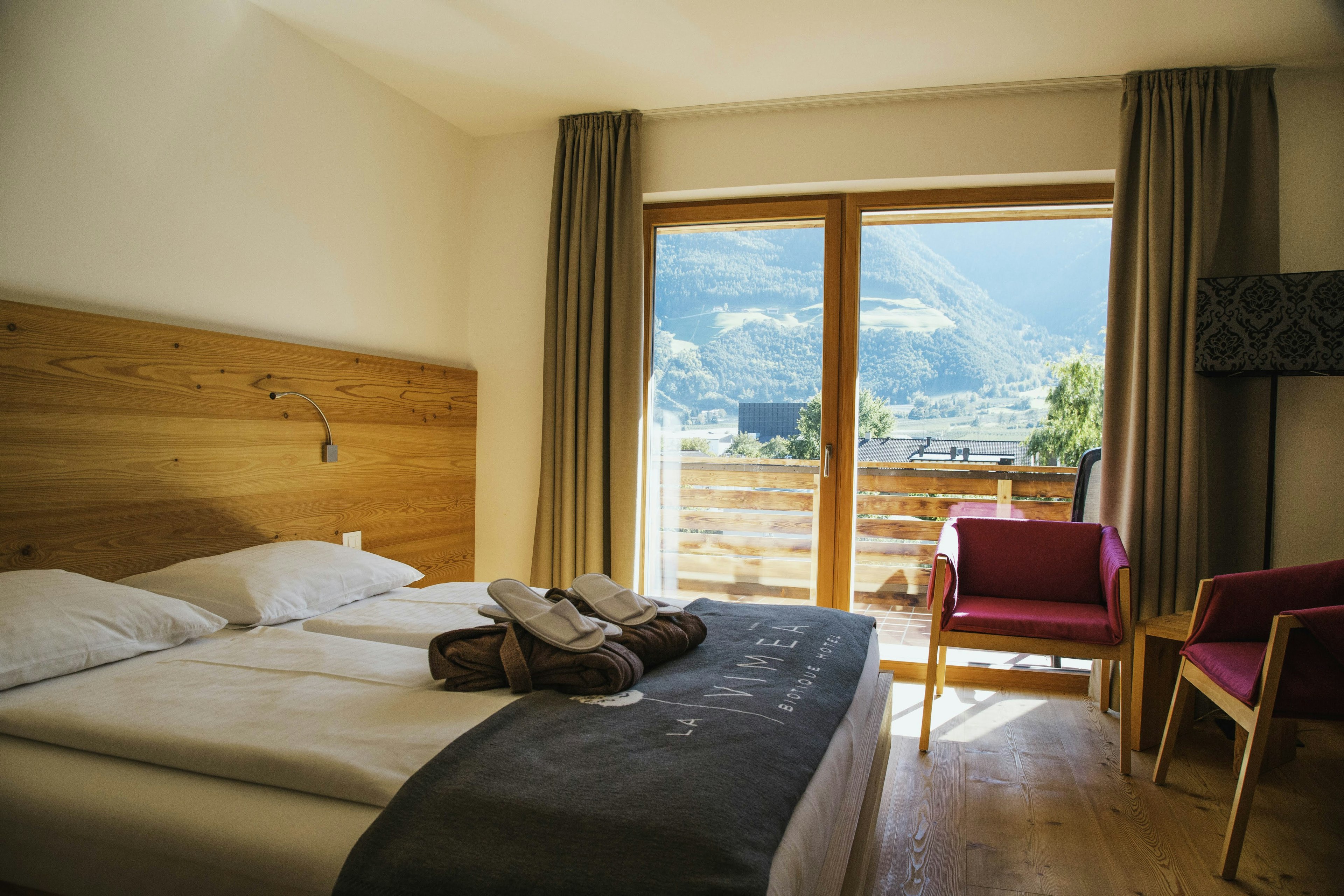 Inside a hotel room with a bed with a wooden backboard and a pair of chairs near a large window. Through the window you can see a large verdant mountain in the distance.