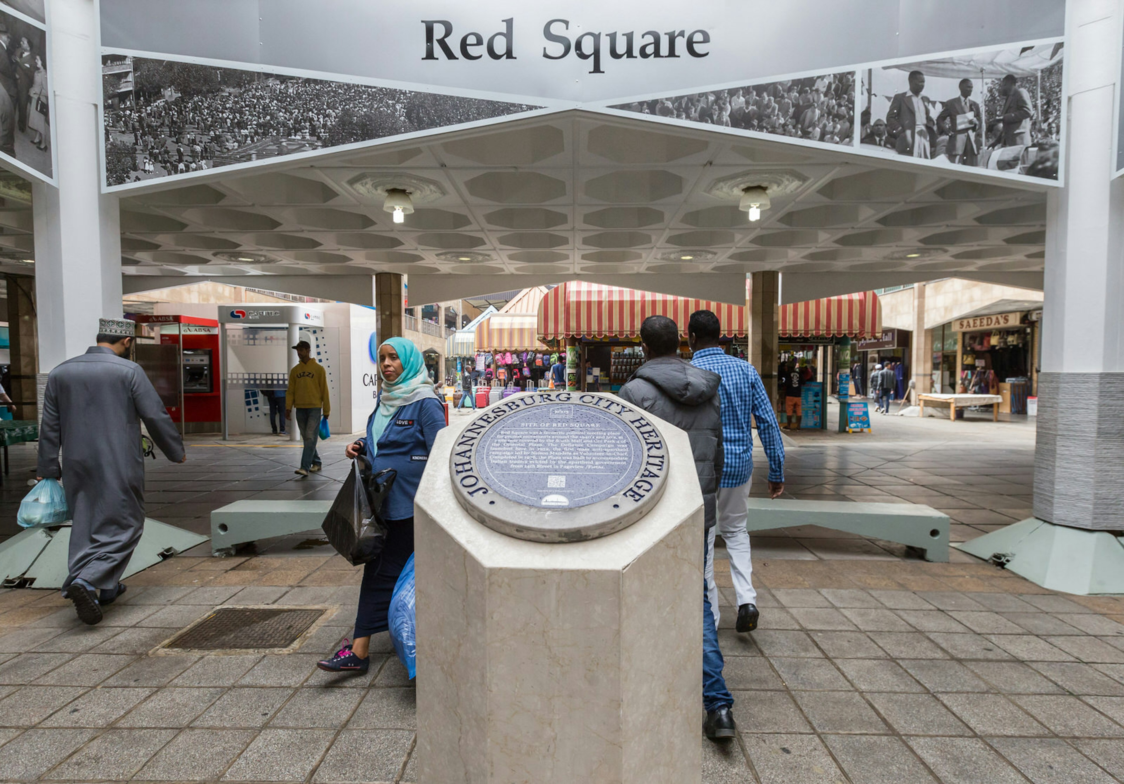 Atop an octagon cement pillar is a plaque entitled 'Johannesburg City Heritage'. Its text speaks of the site of Red Square, which sits in the immediate background of the pillar. People walk about in various styles of dress: a woman in a long skirt and hijab; a man in a long robe and embroidered fez hat; a couple of men in jeans and hoody tops. Fordsburg is one of Johannesburg's best neighbourhoods © Heather Mason / ϰϲʿ¼