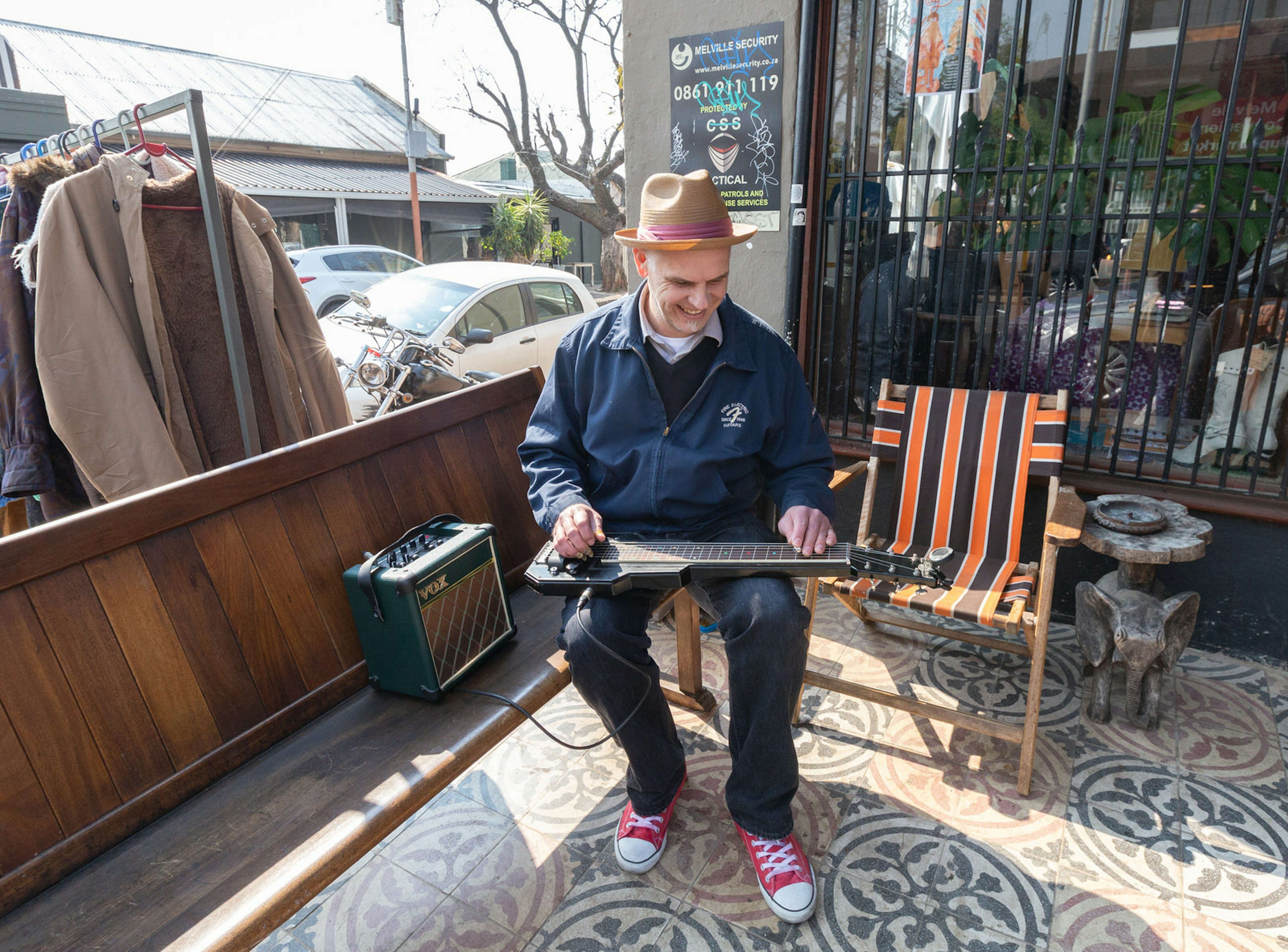 A hip-looking gentleman – in red converse Chuck Taylor shoes, a trilby hat and jeans – sits in a deck chair on the pavement and plays a guitar that is laid flat across his lap. Melville is one of Johannesburg's best neighbourhoods © Heather Mason / ϰϲʿ¼