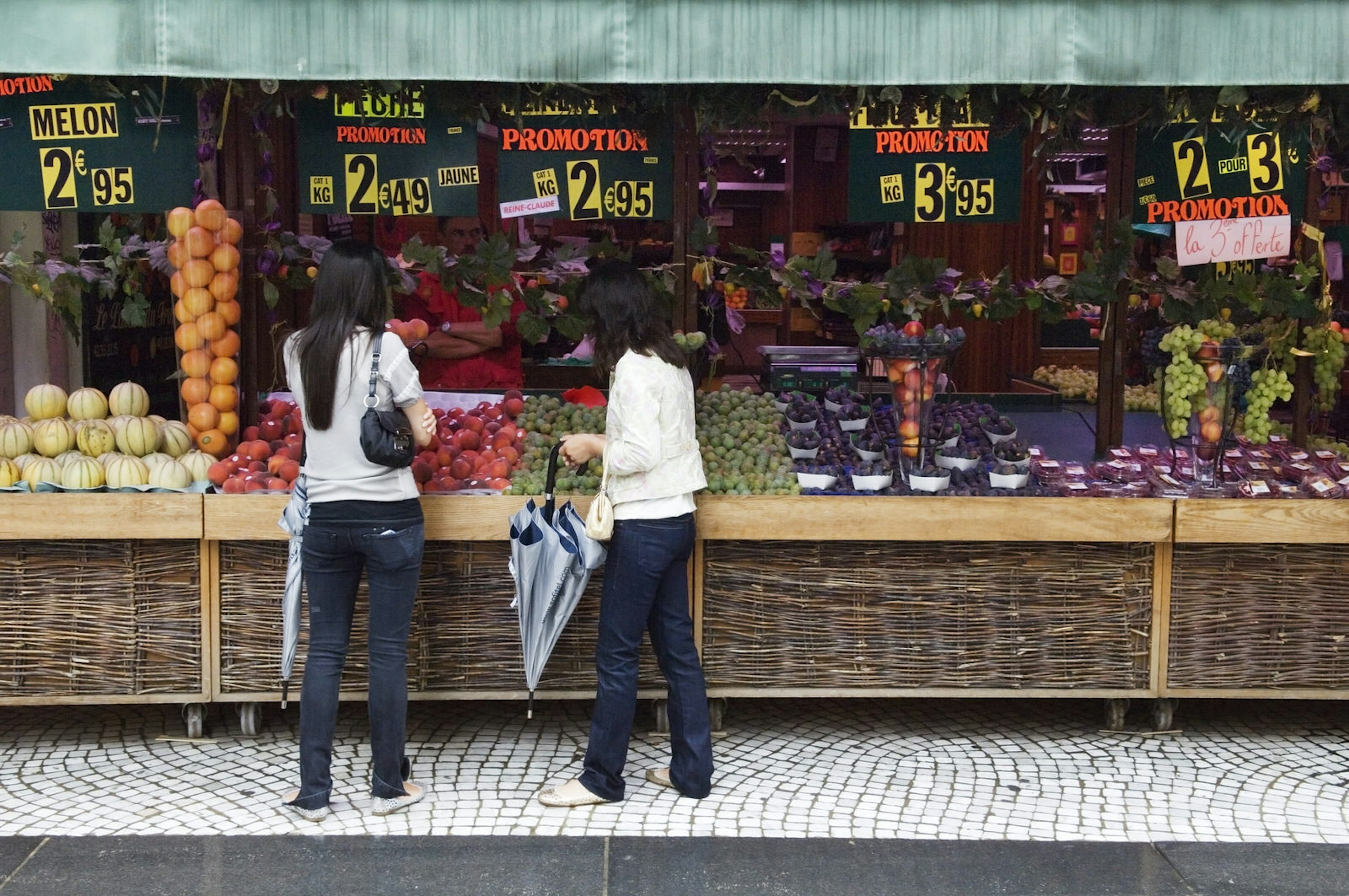 Two people stand in front of a wide range of fruit, displayed below large price signs