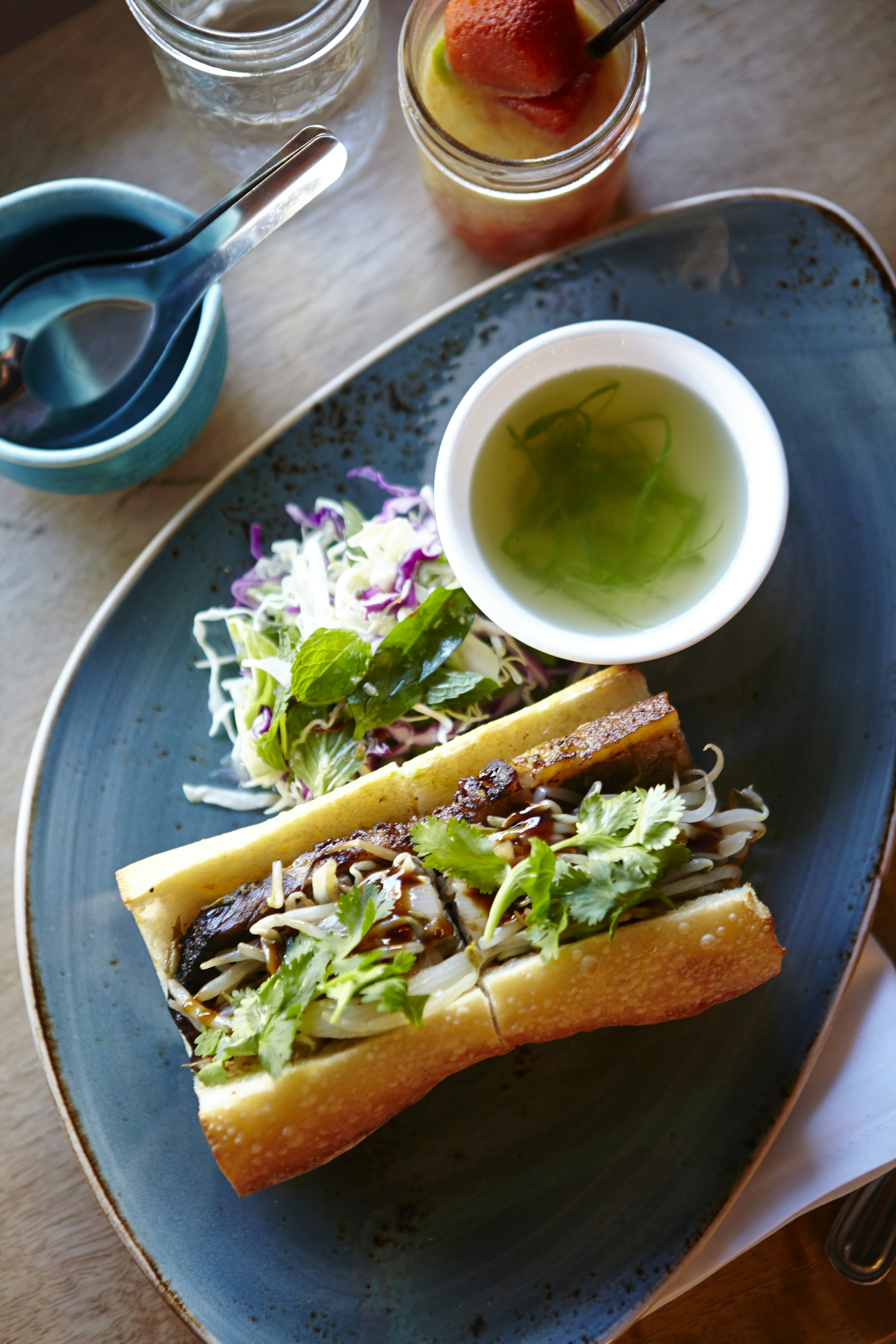 A banh mi sandwich with a side of slaw and a cup of soup are placed on a light blue plate. There are a pair of condiments sitting to the left of the plate; hidden Honolulu