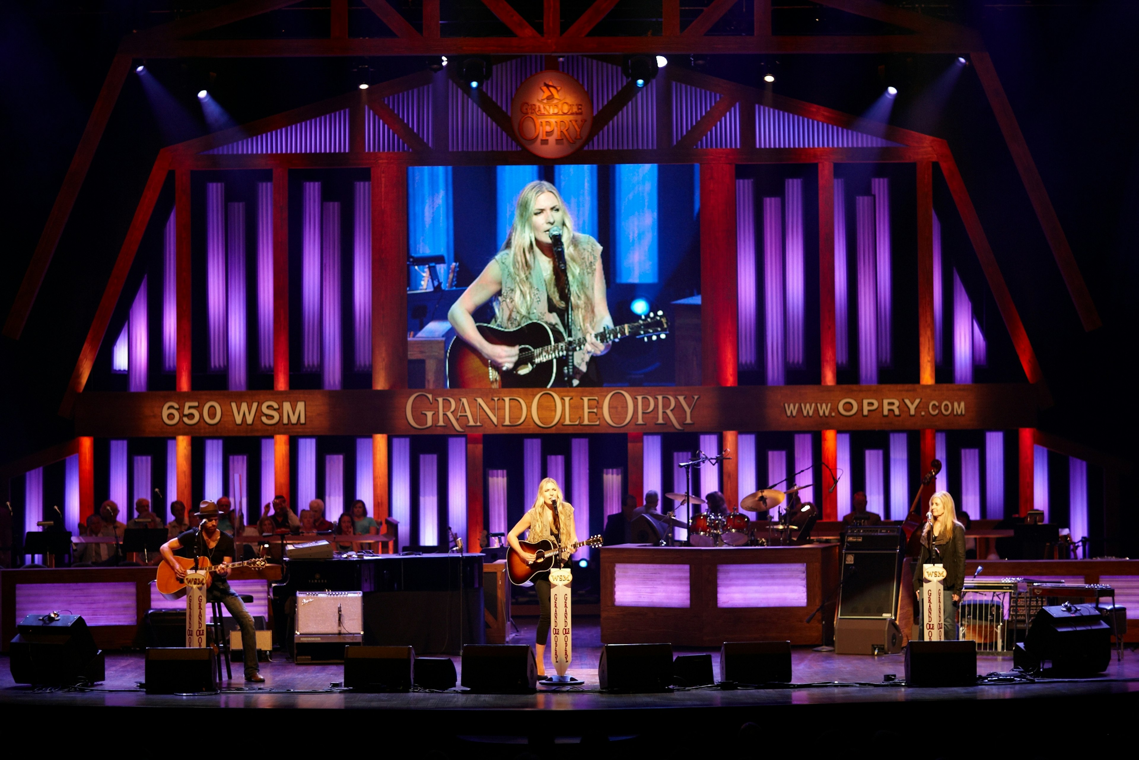 A woman with blond hair and a guitar performs in front of a purple and red backdrop at the Grand Ol Opry in Nashville