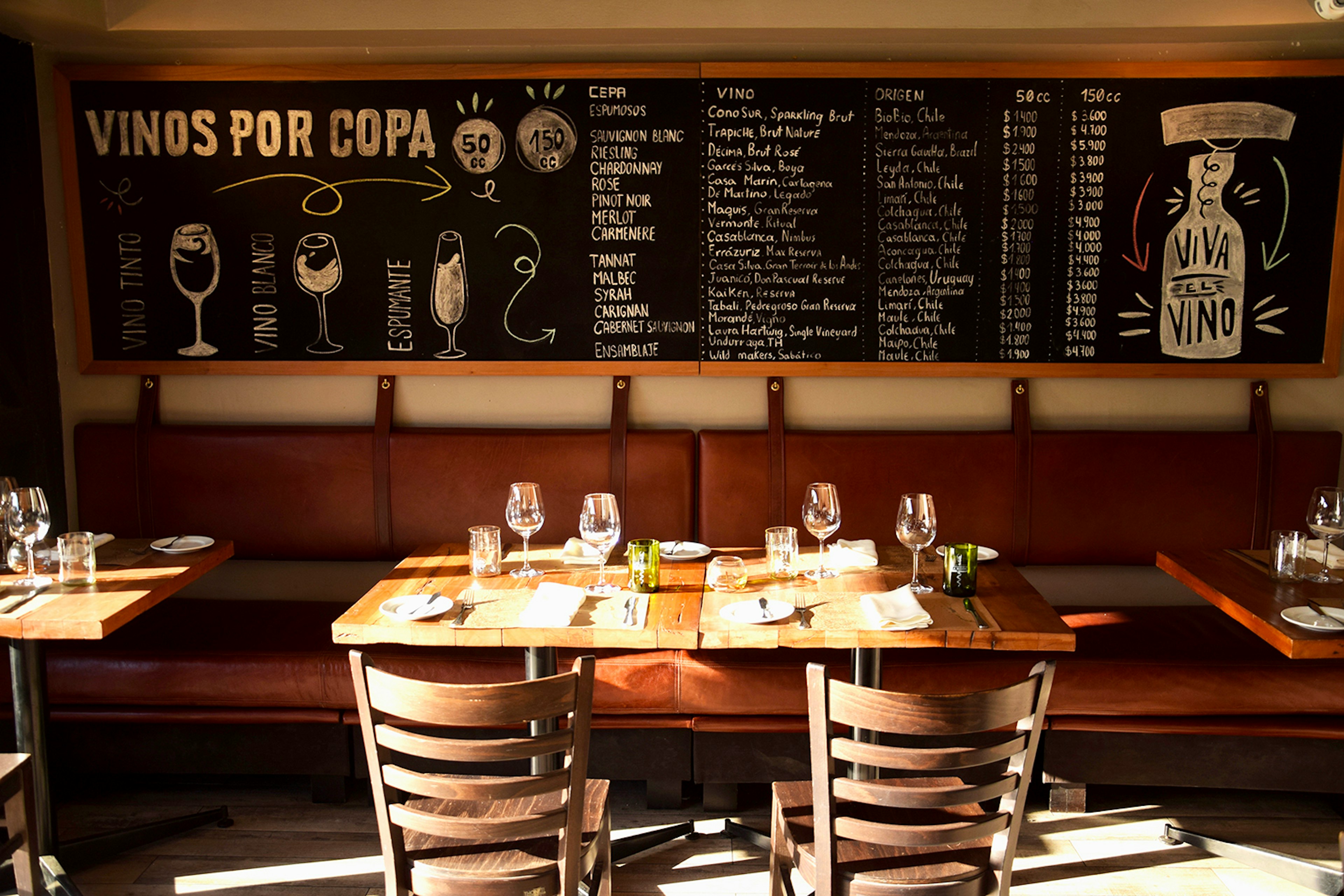 An image of an empty table set with wineglasses and hit with sun, with a chalk board listing wines in the background