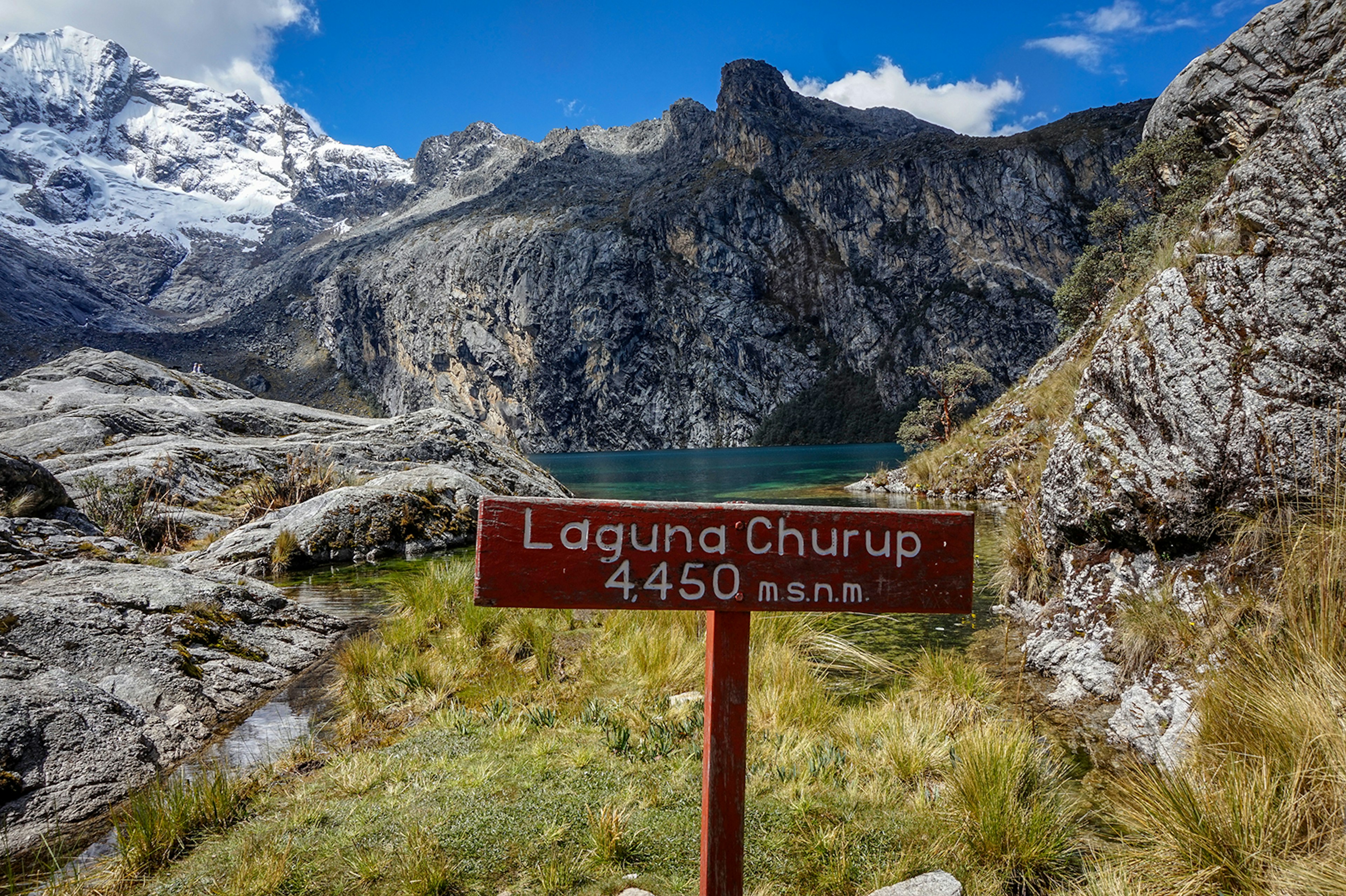 A clear mountain lake surrounded by rocky, snowy peaks, with an altitude sign for Laguna Churup in the foreground © Brendan Sainsbury / Lonely Planet