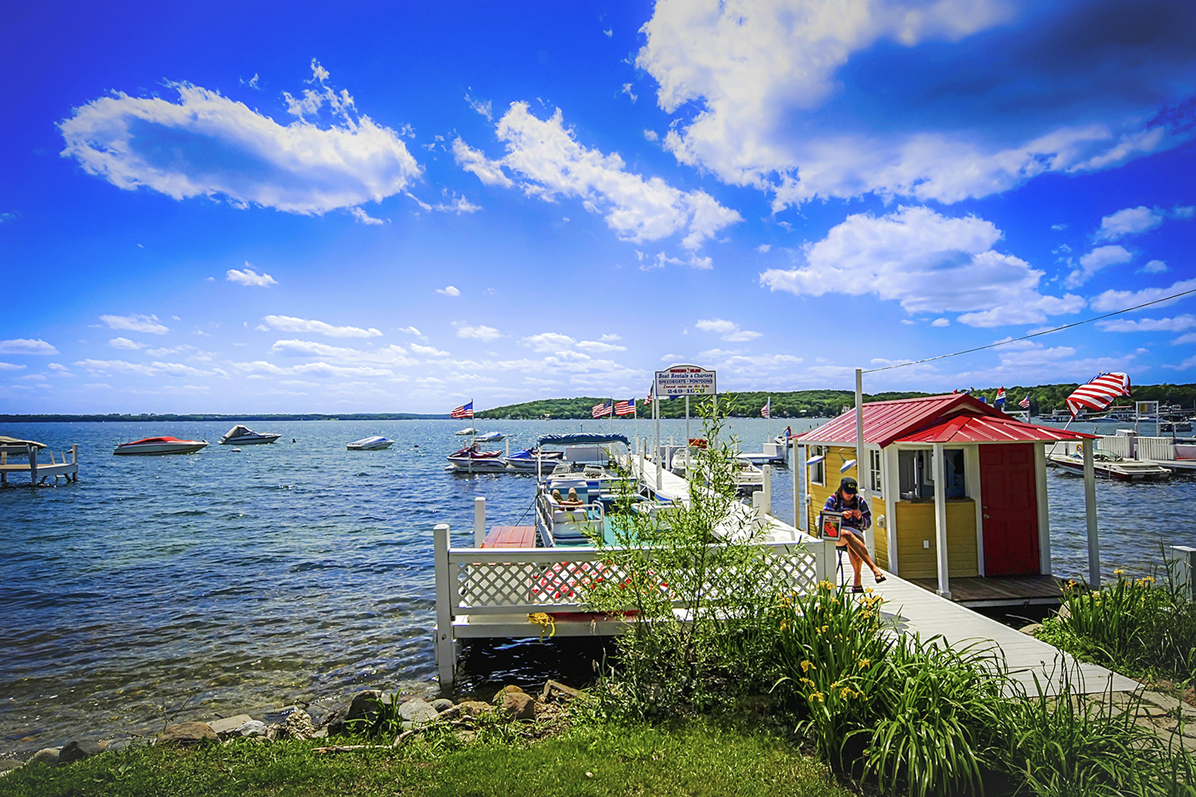 An image of a small lakeside pier next to a floating building with a red roof, with some boats floating on the water in the background at Lake Geneva, Wisconsin