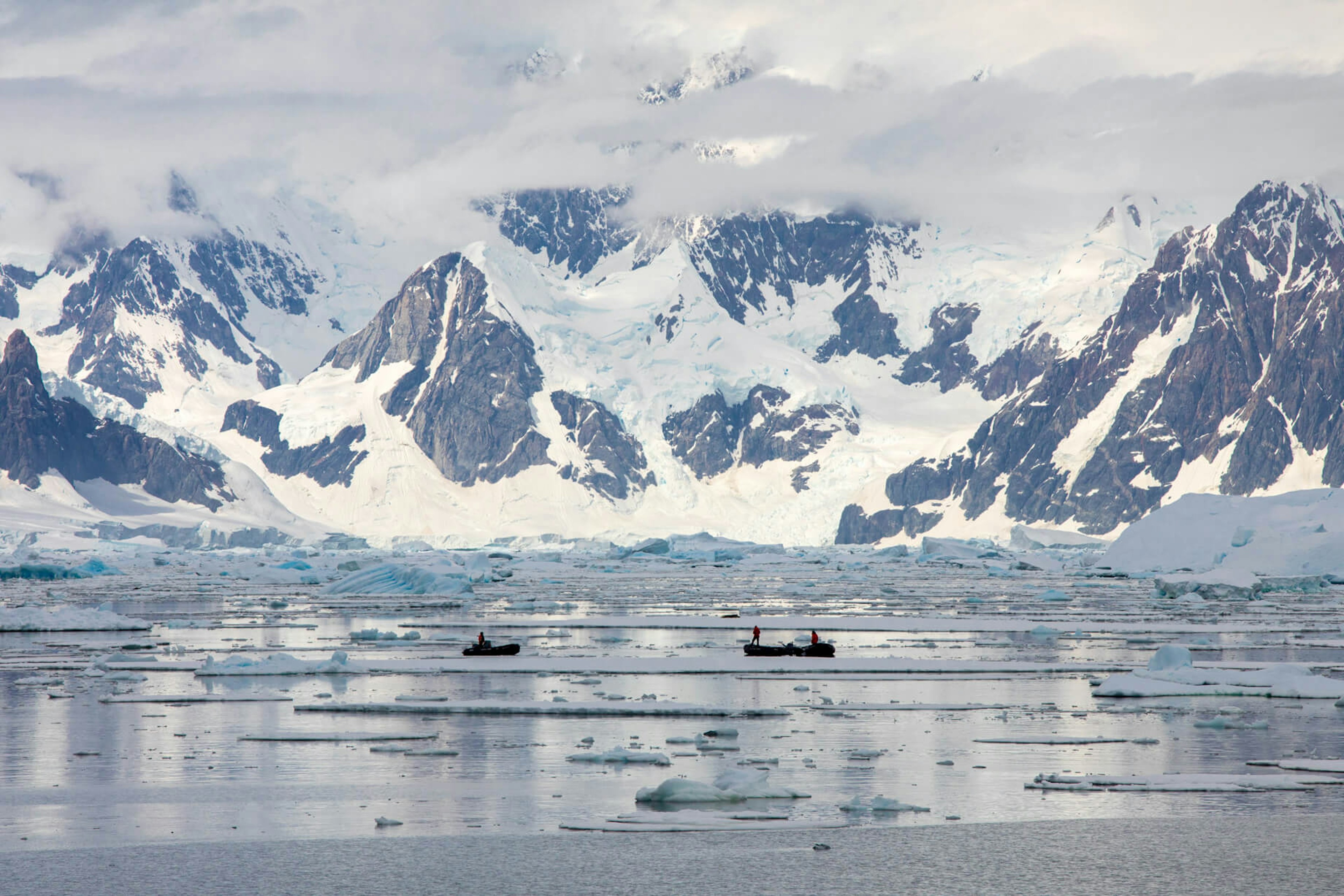 Two small boats navigate the fjord. They are dwarfed by huge mountains and glaciers in the background.