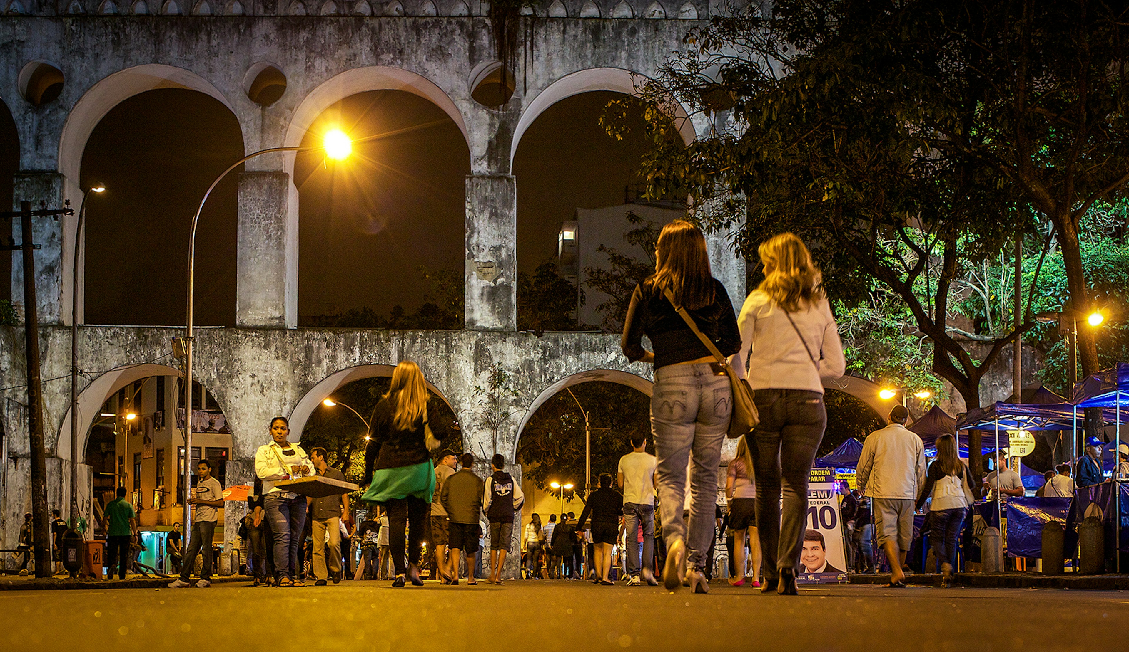 Two women in the foreground, with their backs to the camera, walk towards a crowd of people and a large whitewashed aqueduct