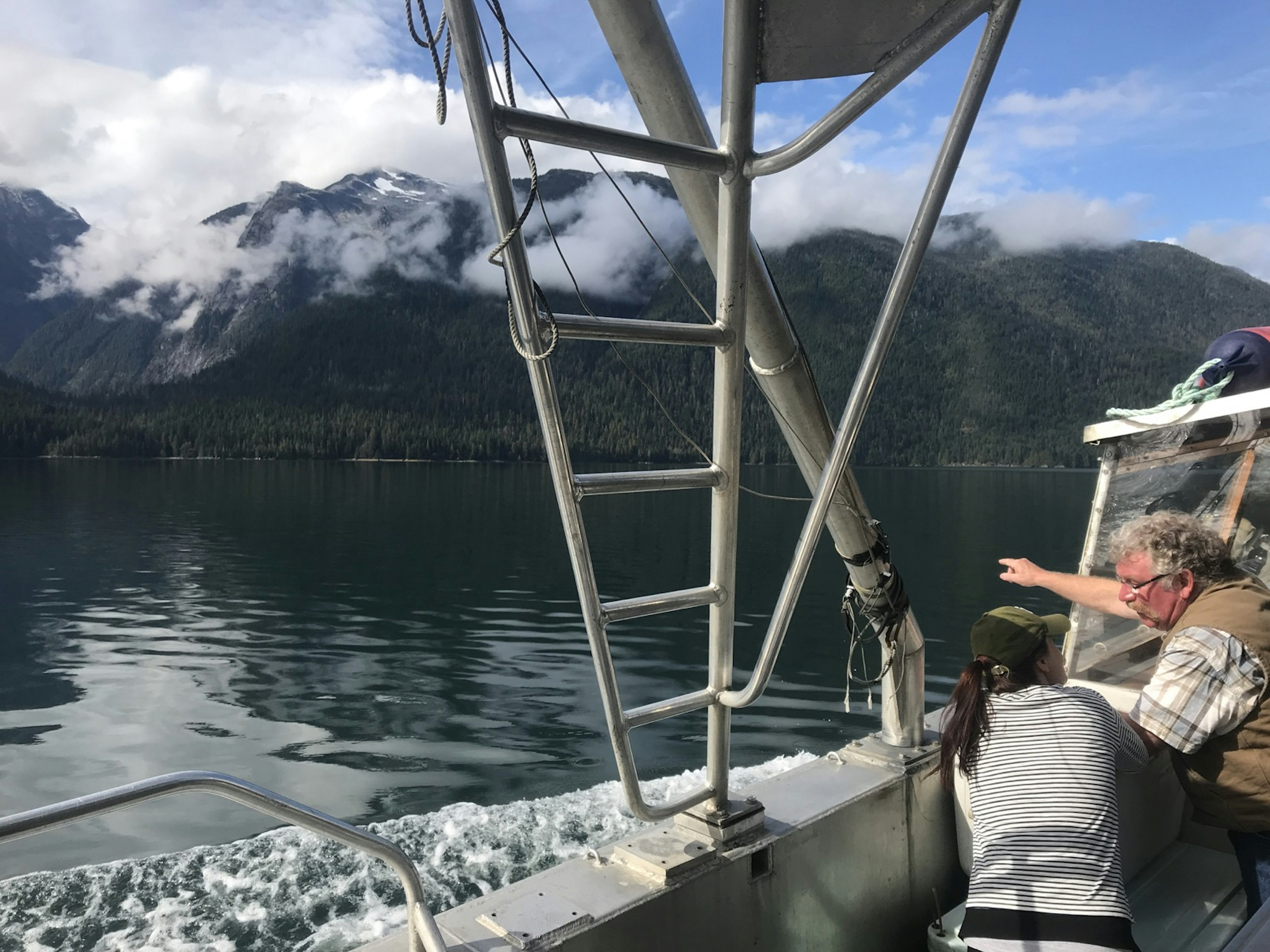 Mist clings to a forested mountain as a man with a giant handlebar mustache gestures from the deck of a boat