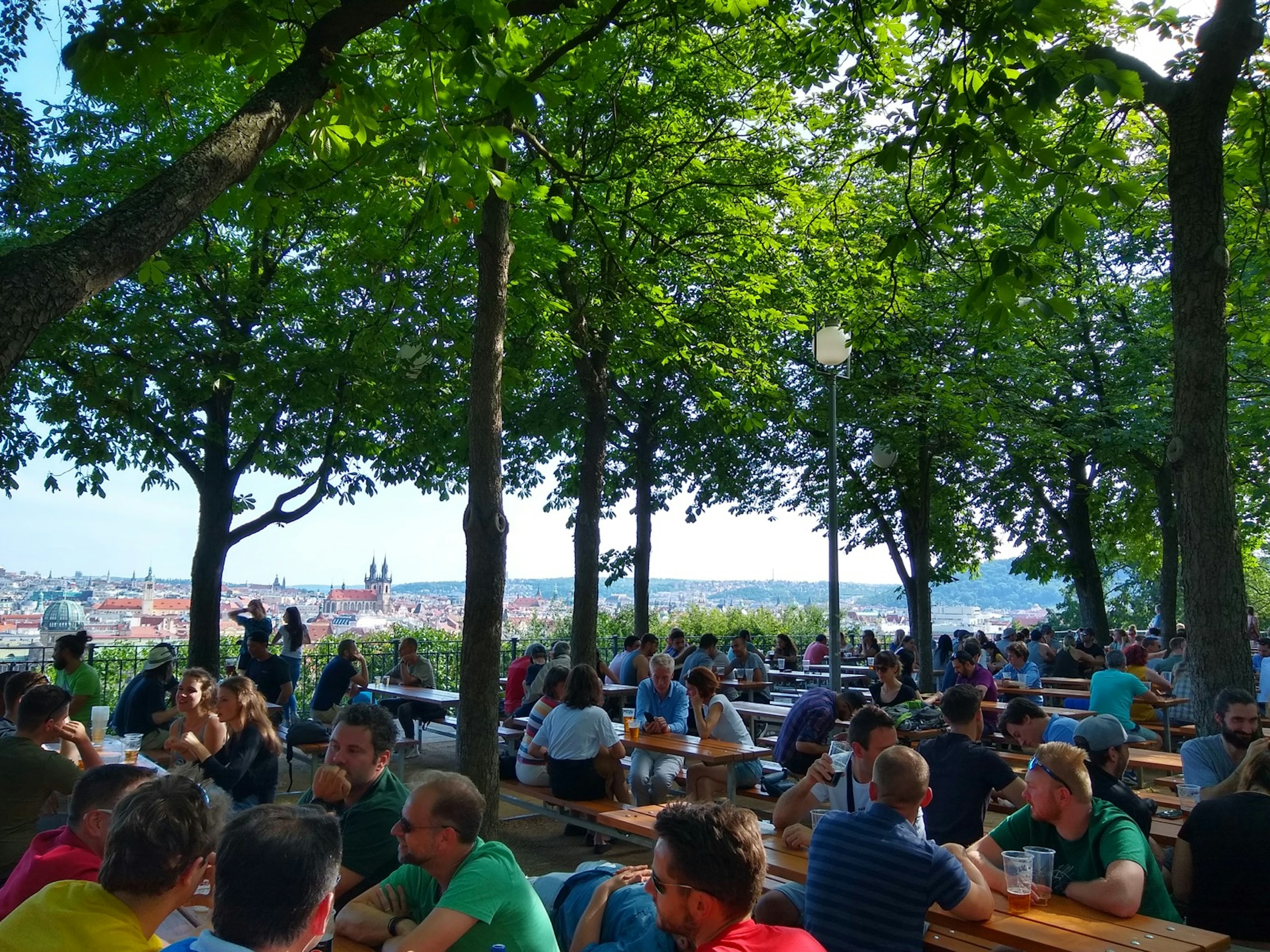 Rows of tables full of beer-drinking people are arranged under a shady canopy of trees on a bluff that overlooks the city of Prague