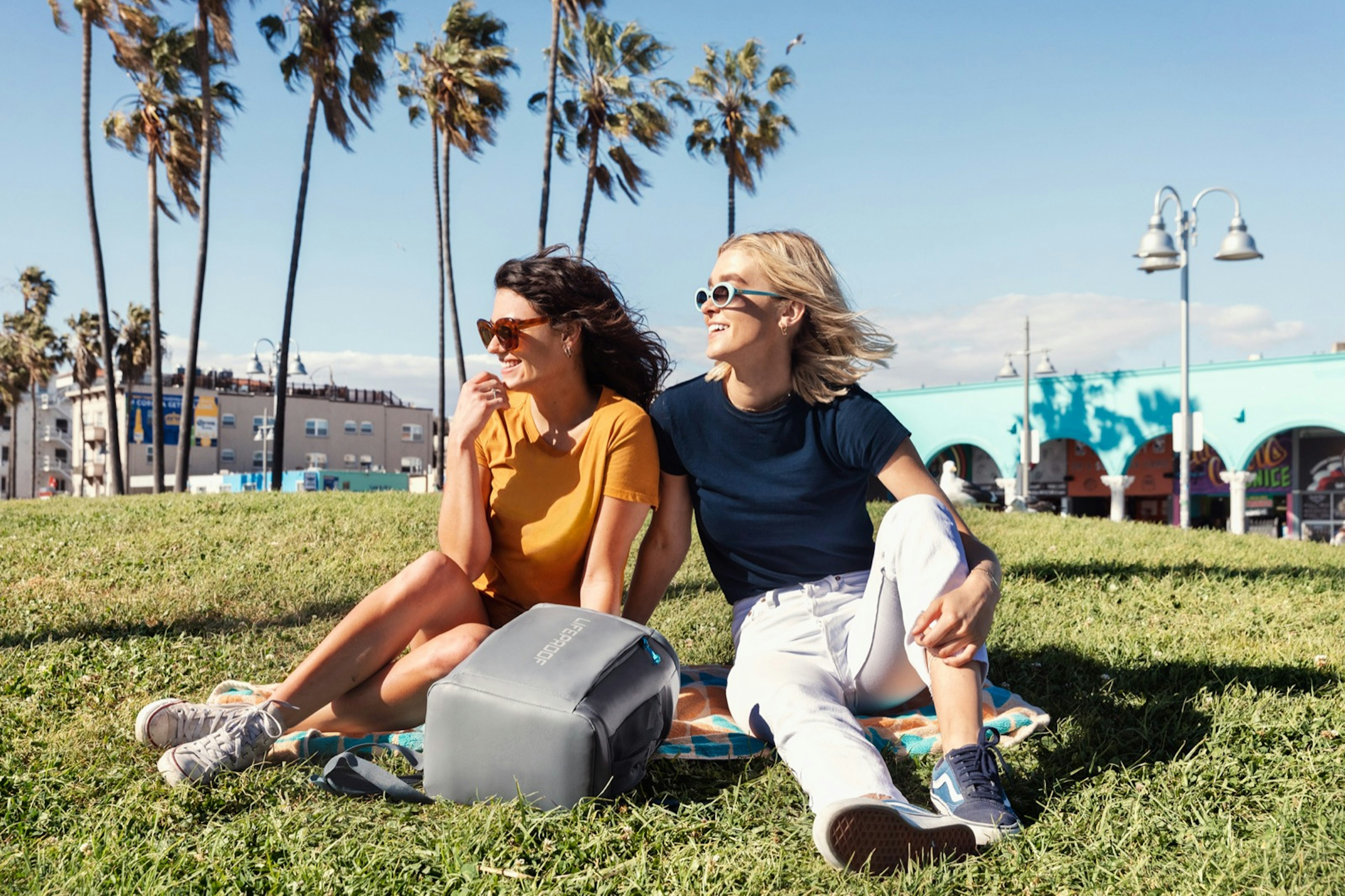 Two women in a sunny park with a backpack cooler on a beach towels, with palm trees in the distance; beach gear