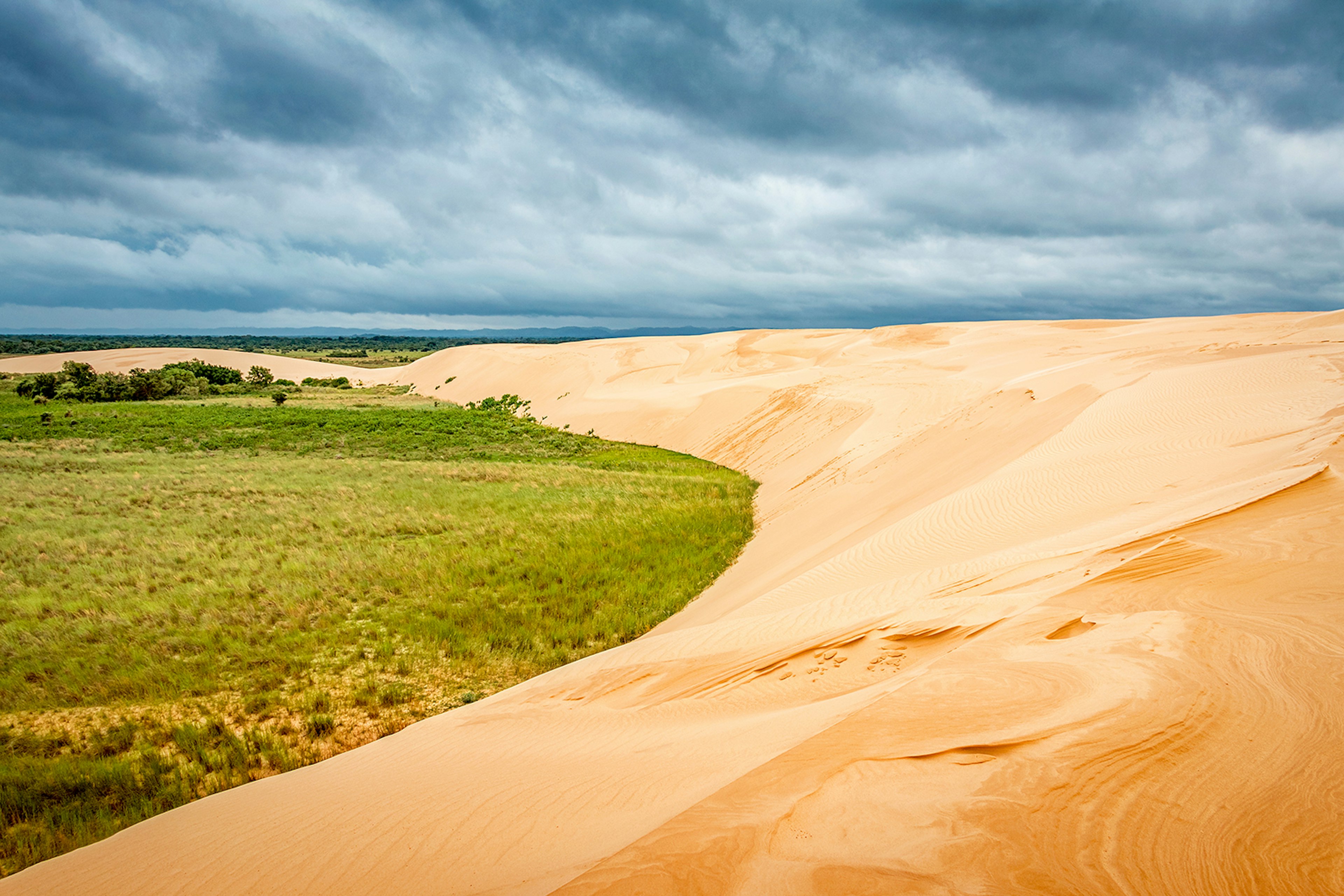 A large arc of golden sand sits to the right of a green field in Parque Lomas de Arena, Bolivia