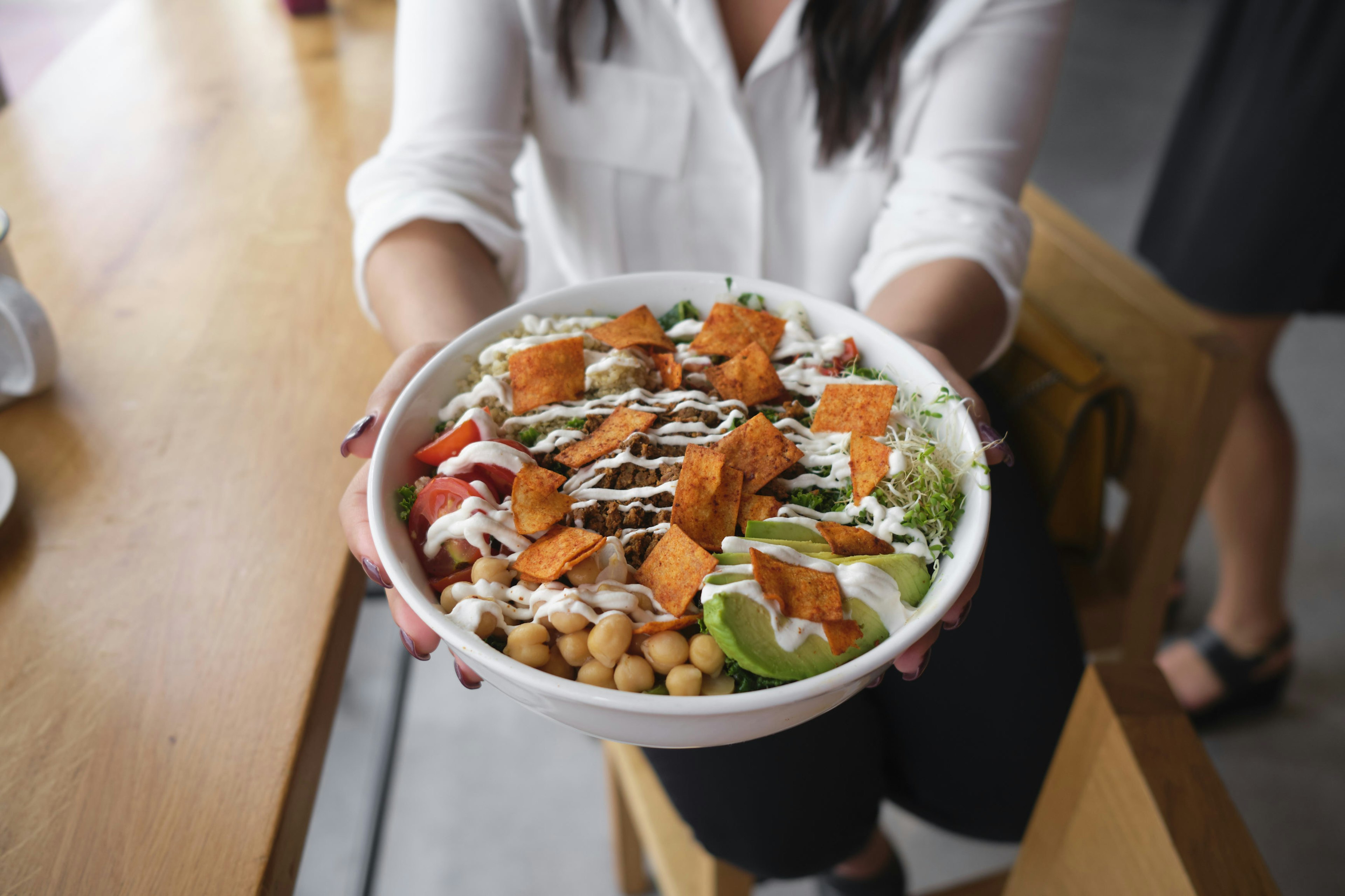 A person holds a bowl filled with chick peas, tomatoes, avocado and other veggies is topped with a white sauce; vegan restaurant miami