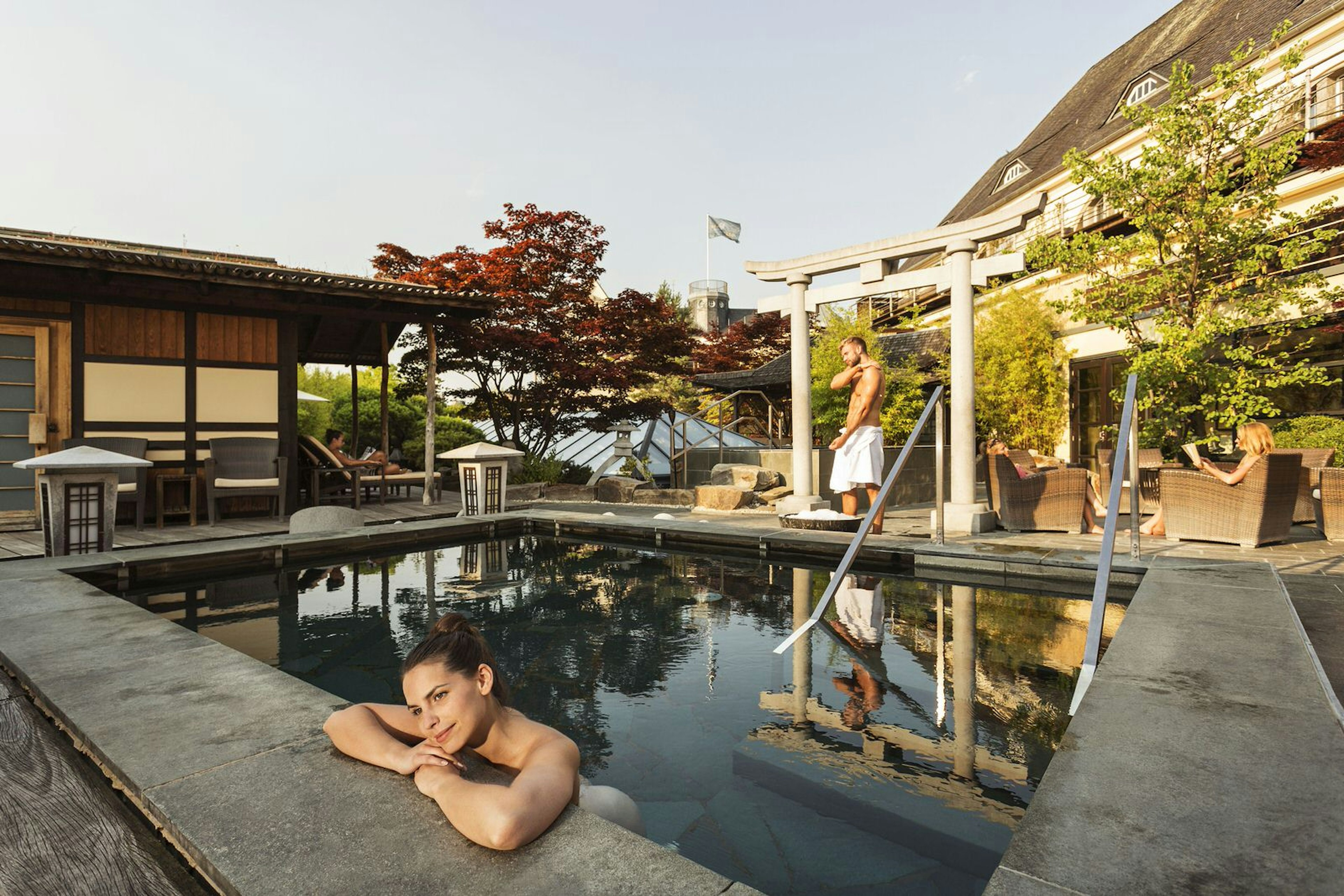 People relaxing in an outdoor pool at Neptunbad, Cologne. The pool is surrounded by a zen garden, featuring trees and gateways