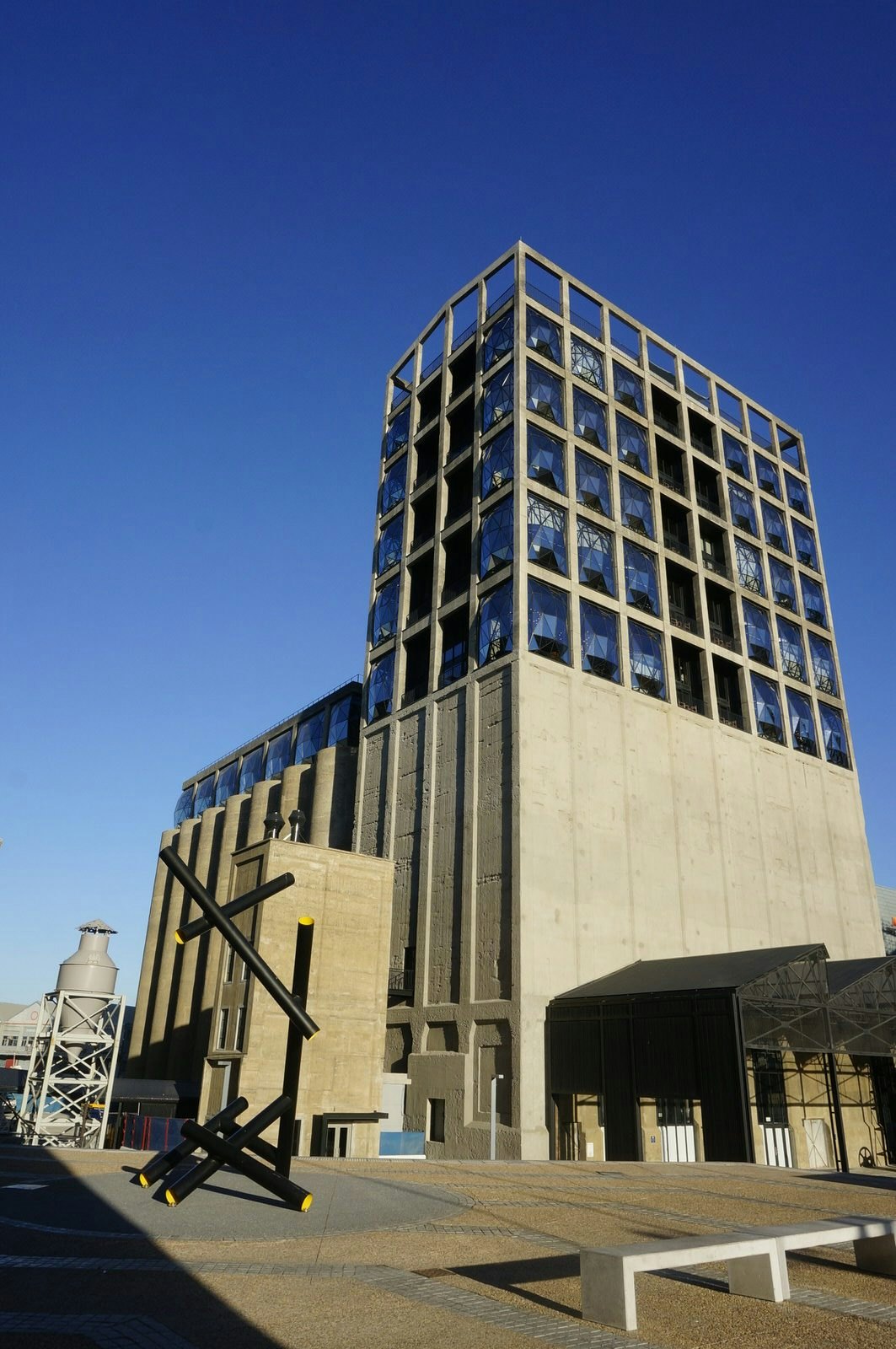 Housing African art and standing in front of a brilliant blue sky is the grey concrete facade of the Zeitz MOCAA. Comprised of two parts, one being a tall tower with lots of geodesic windows in a grid pattern on its upper half (lower half being solid concete), the other being the grouped silos themselves, which are about half the height of the tower © Monica Suma / Lonely Planet