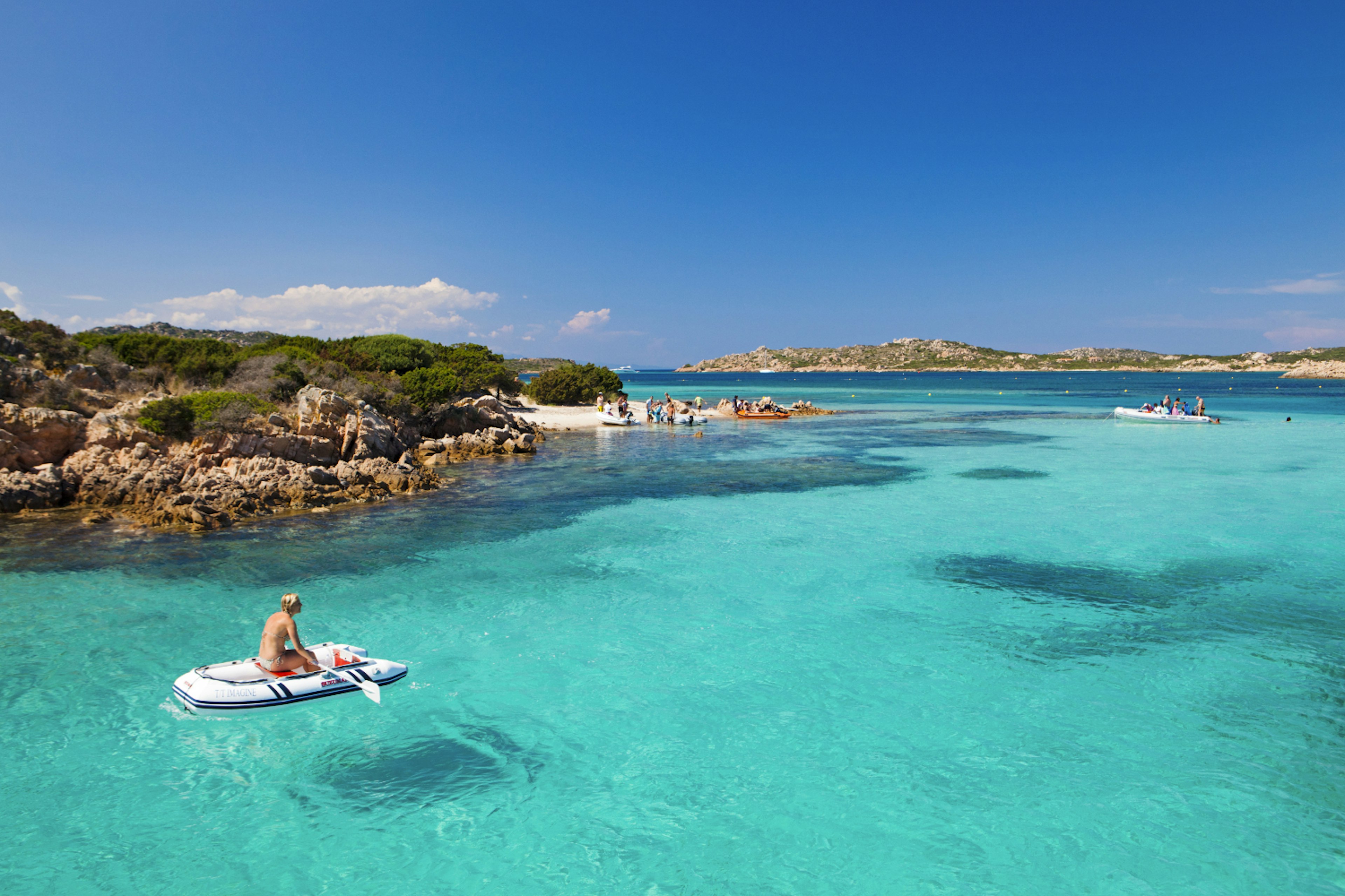 A person sits in a small inflatable boat in the light blue waters of the Isola Maddelena archipelago, Sardinia.