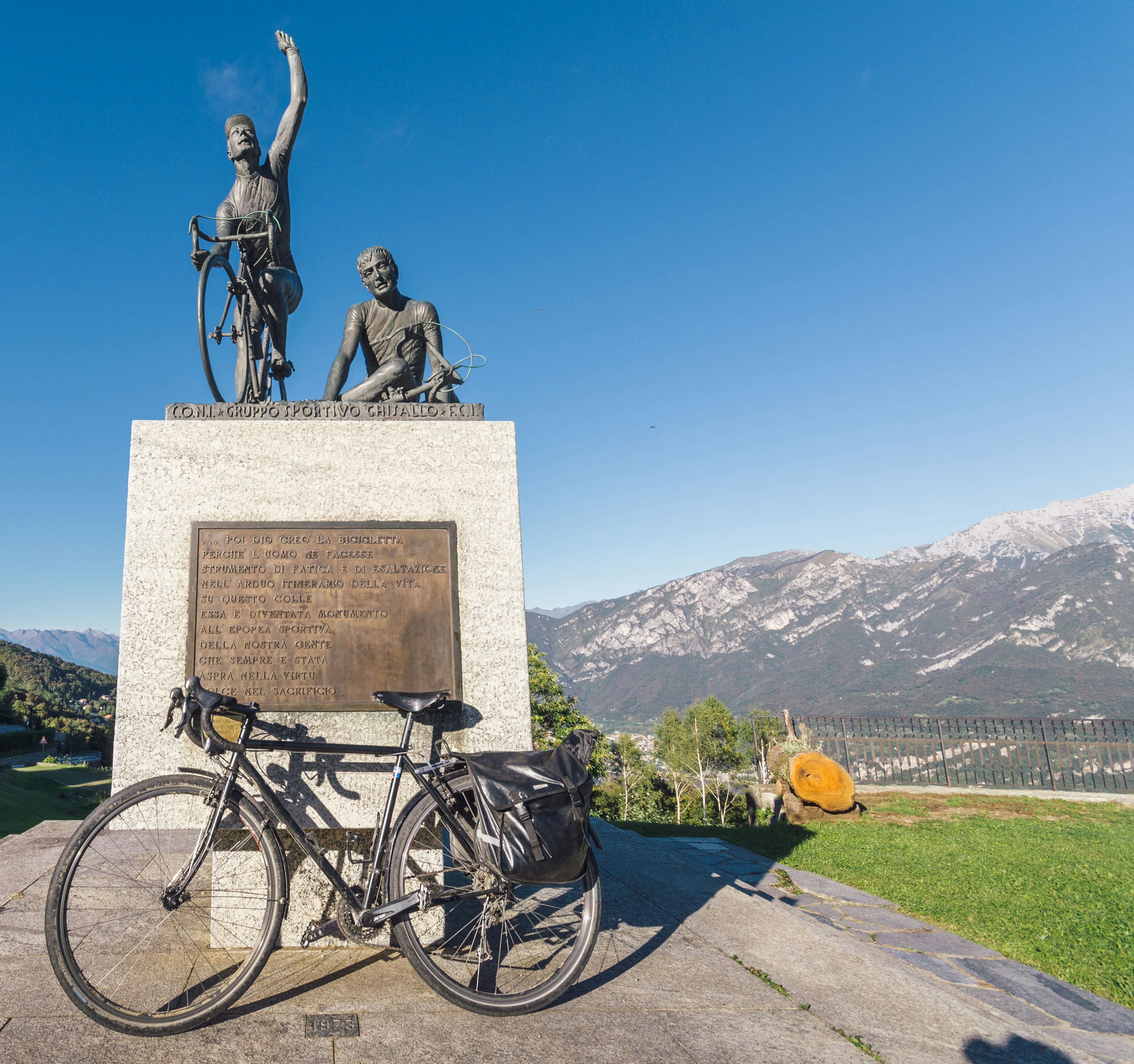 A bike sits in front of a tall monument in memory of cyclists to the Madonna del Ghisallo sits in front of a church.