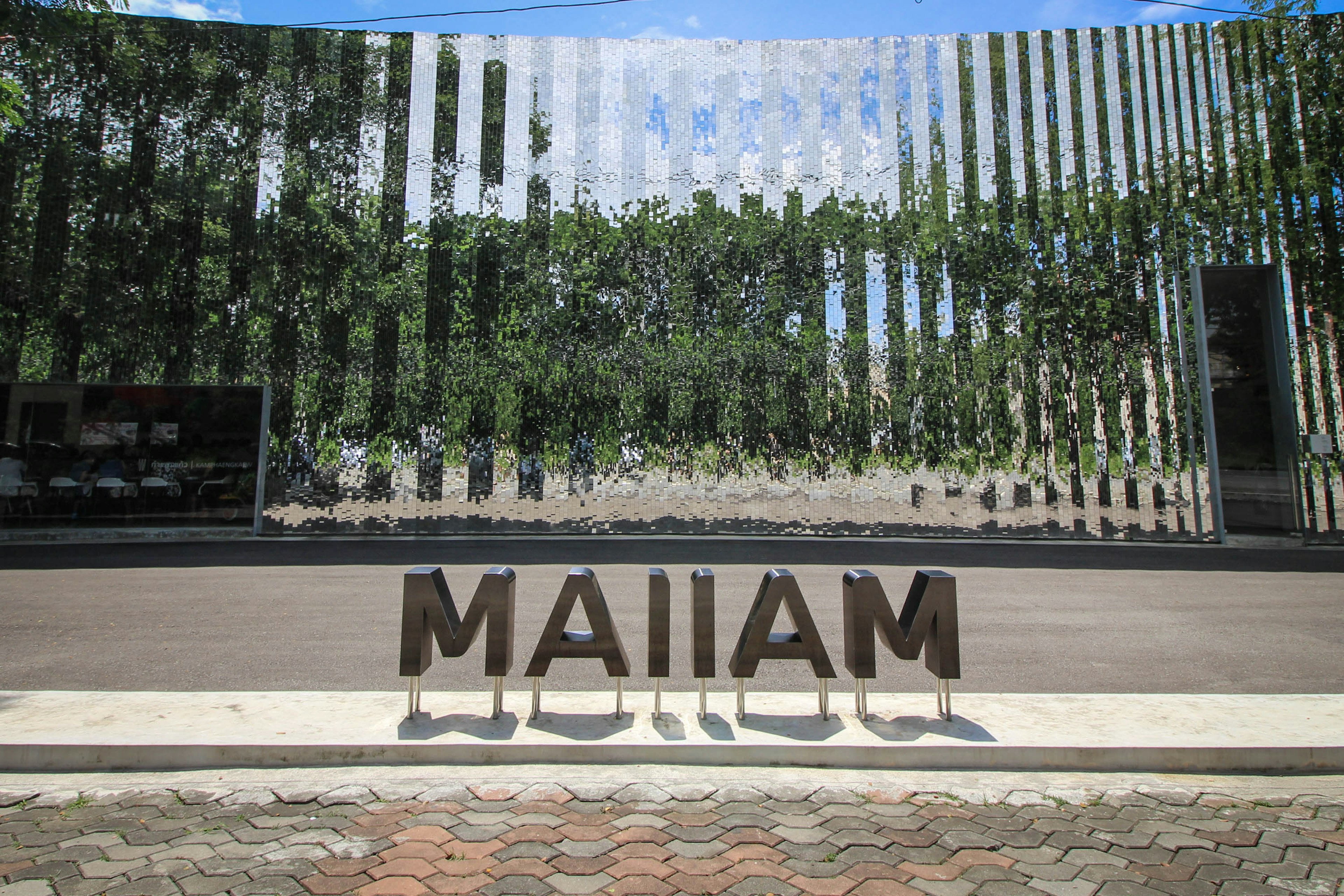 Exterior of the Maiiam Contemporary Art Museum in Chiang Mai. The silver building causes a semi-mirror effect, reflecting the greenery in front of it, while the word 'Maiiam' is spelled in big letters by the entrance.