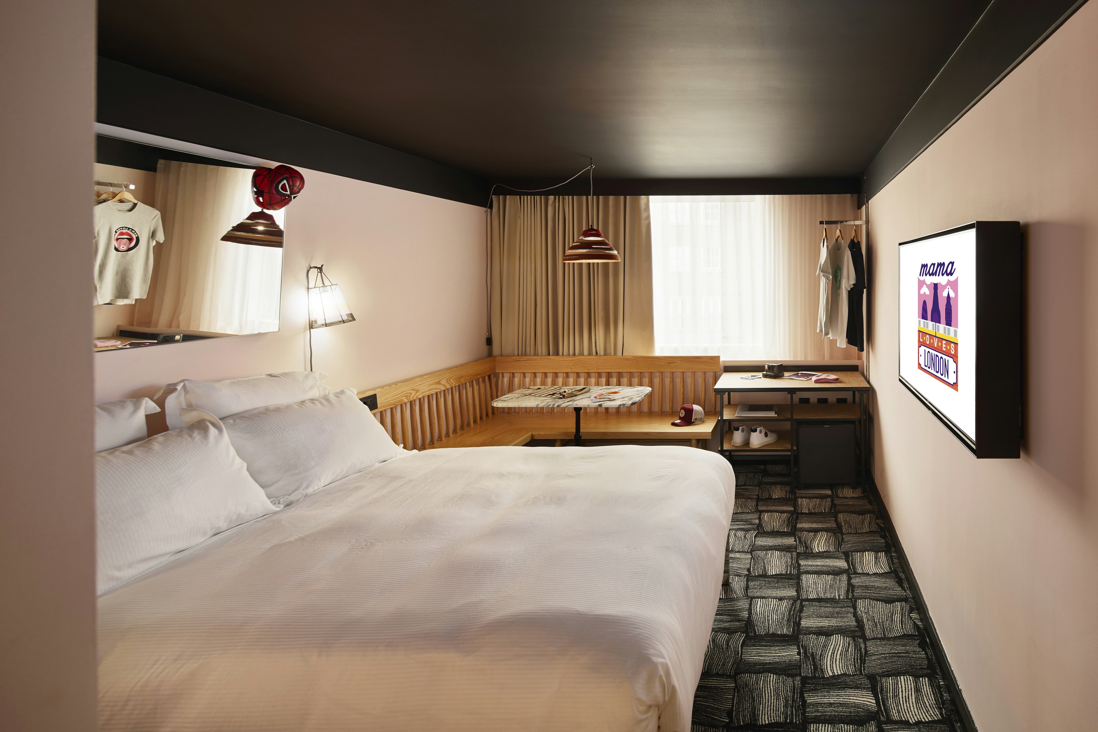 A room at Mama Shelter London, with a neatly made bed with a mirror behind it and bordered by a wrap around bench and table with small shelves.