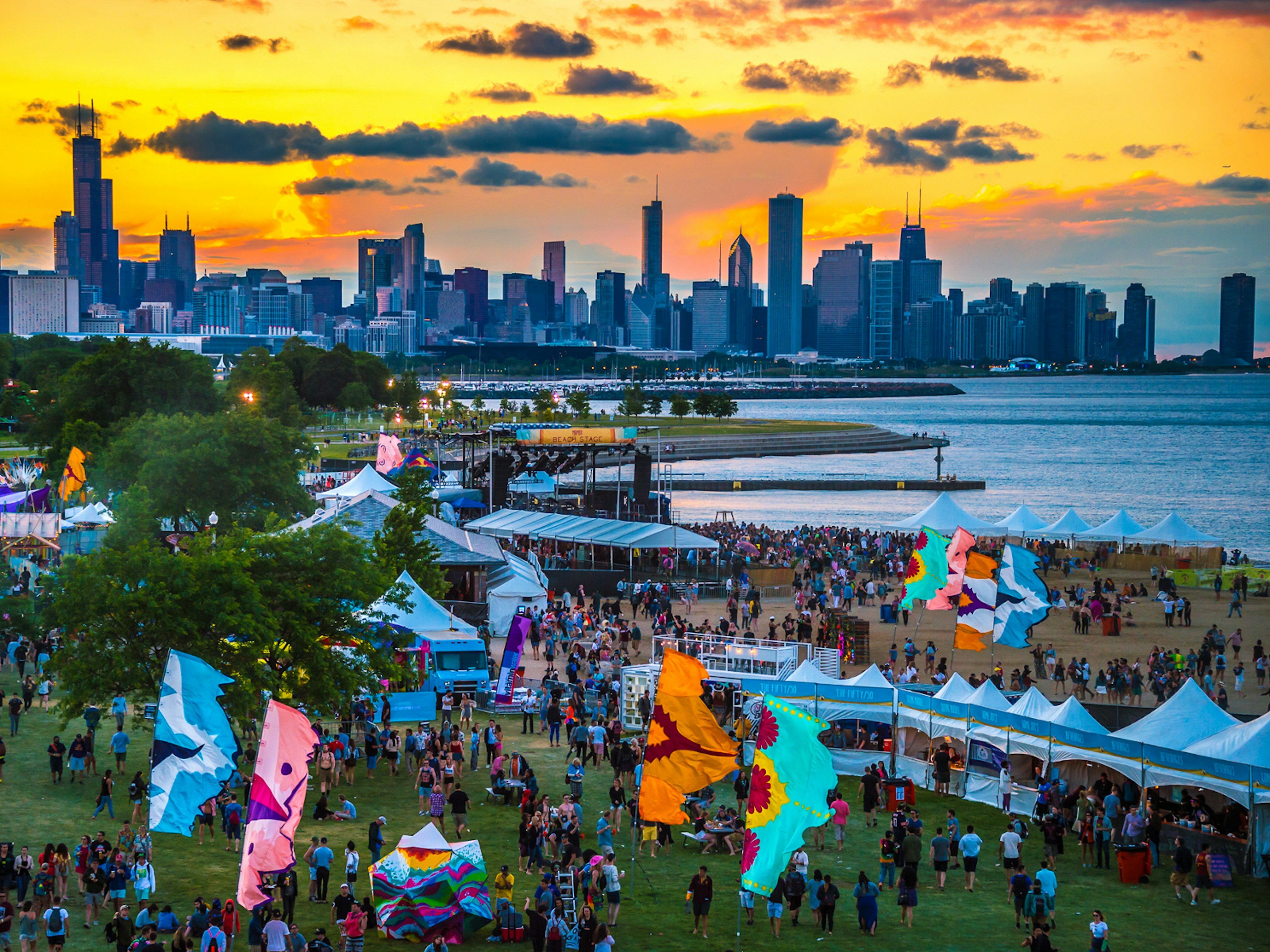 Alternative festivals - Mamby festival on the beach, at dusk with the city skyline in the background. Colourful vertical flags fly around the festival grounds, alongside large white marquees. The background is lit by a vivid yellow and orange sunset behind sparse clouds.