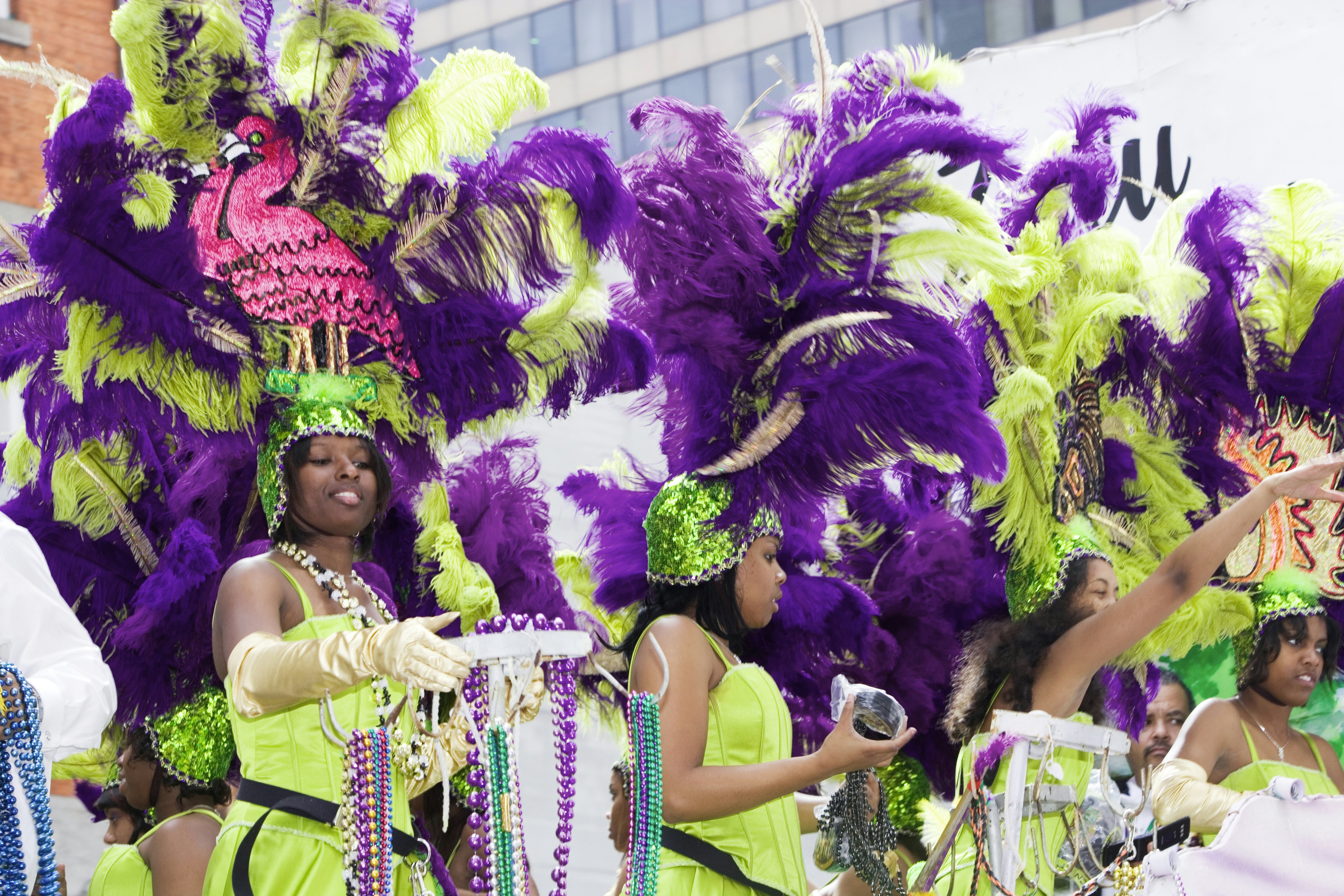 A group of women dressed in lime green dresses and green and purple feather and sequined headdresses throw beads to the crowd during Mardi Gras.