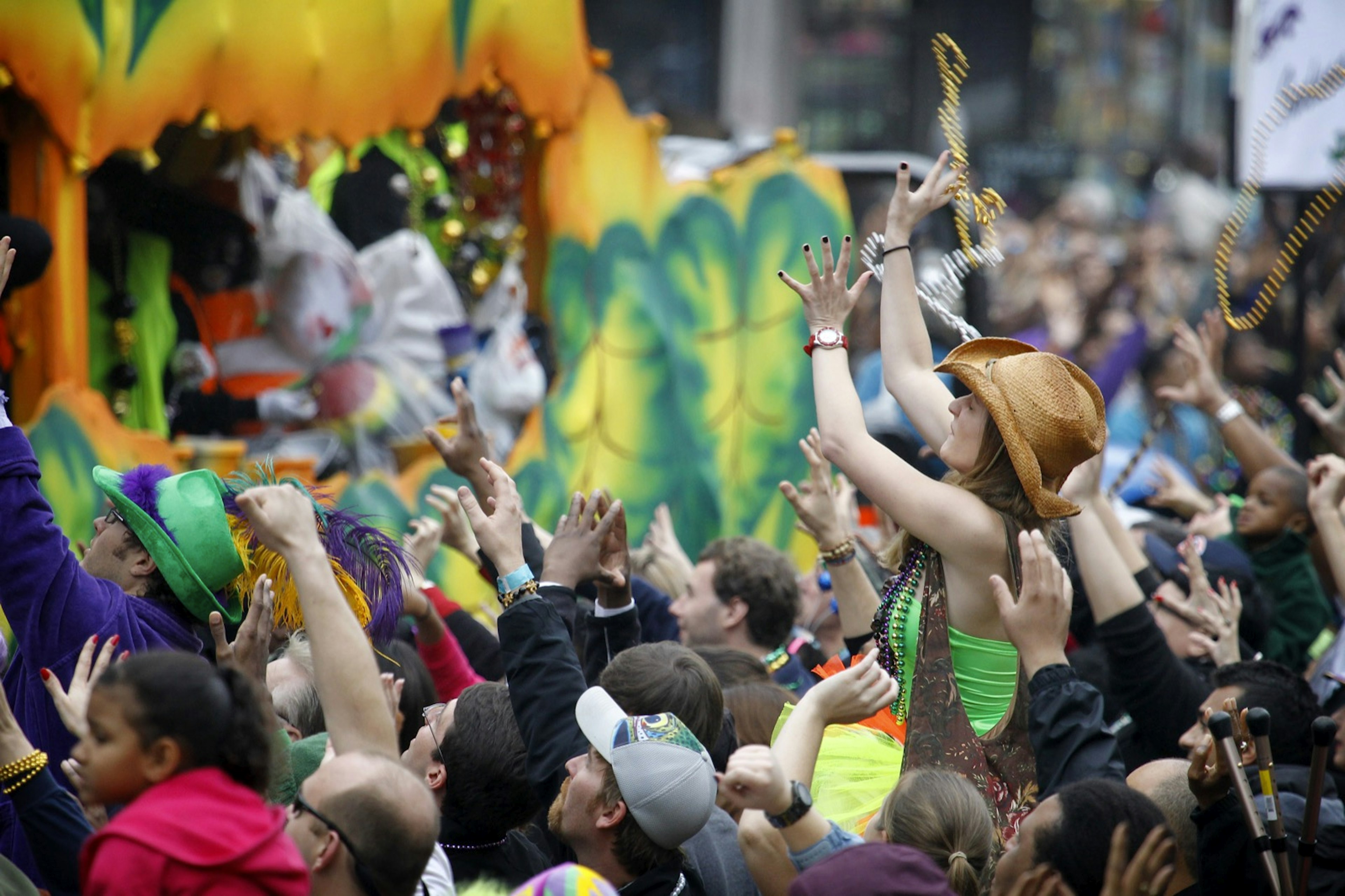 A woman in a cowboy hat sits on someone's shoulders as she tries to catch some beads being thrown from a float at Mardi Gras in New Orleans