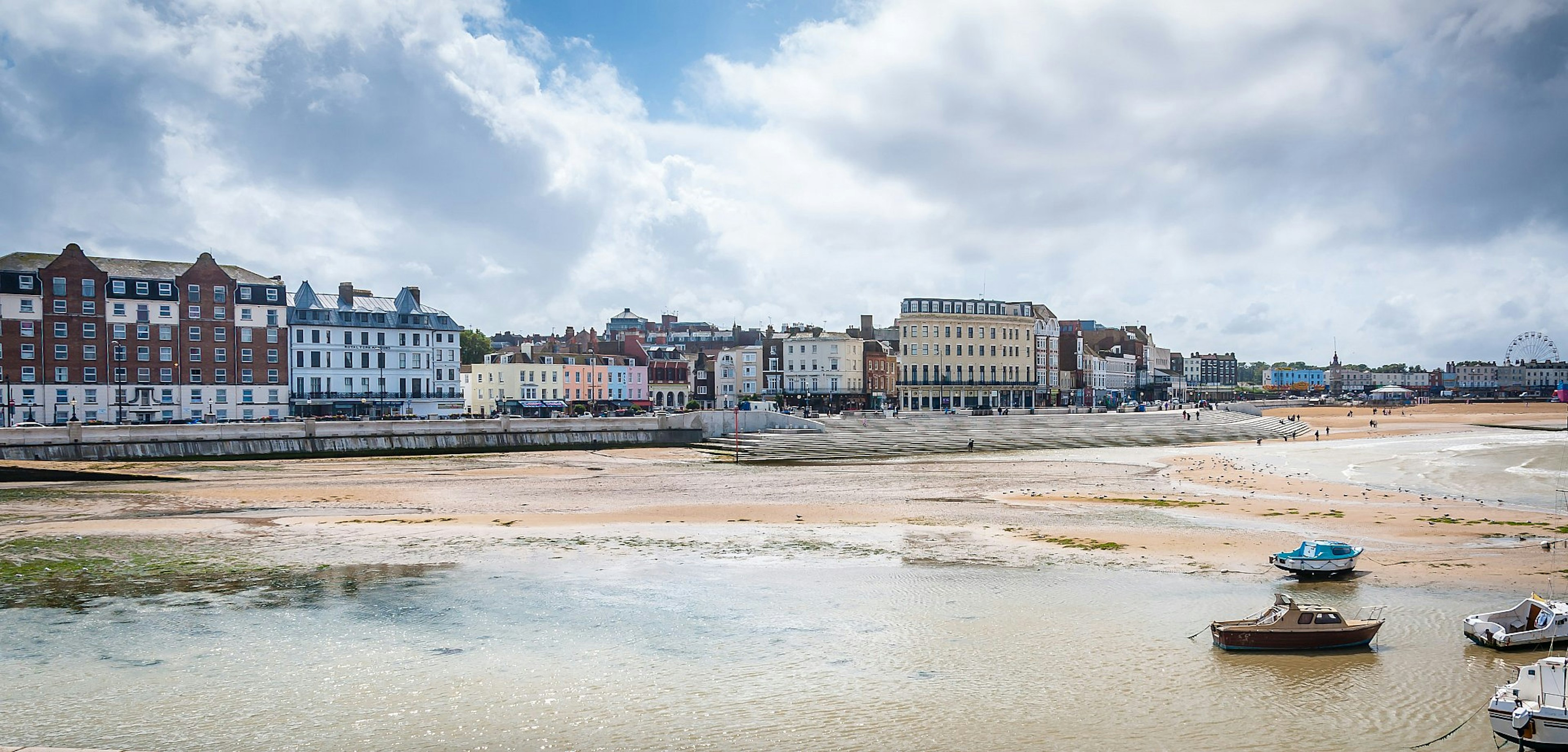 Boats in Margate harbour at low tide, with buildings along the seafront and a Ferris wheel visible in the distance.